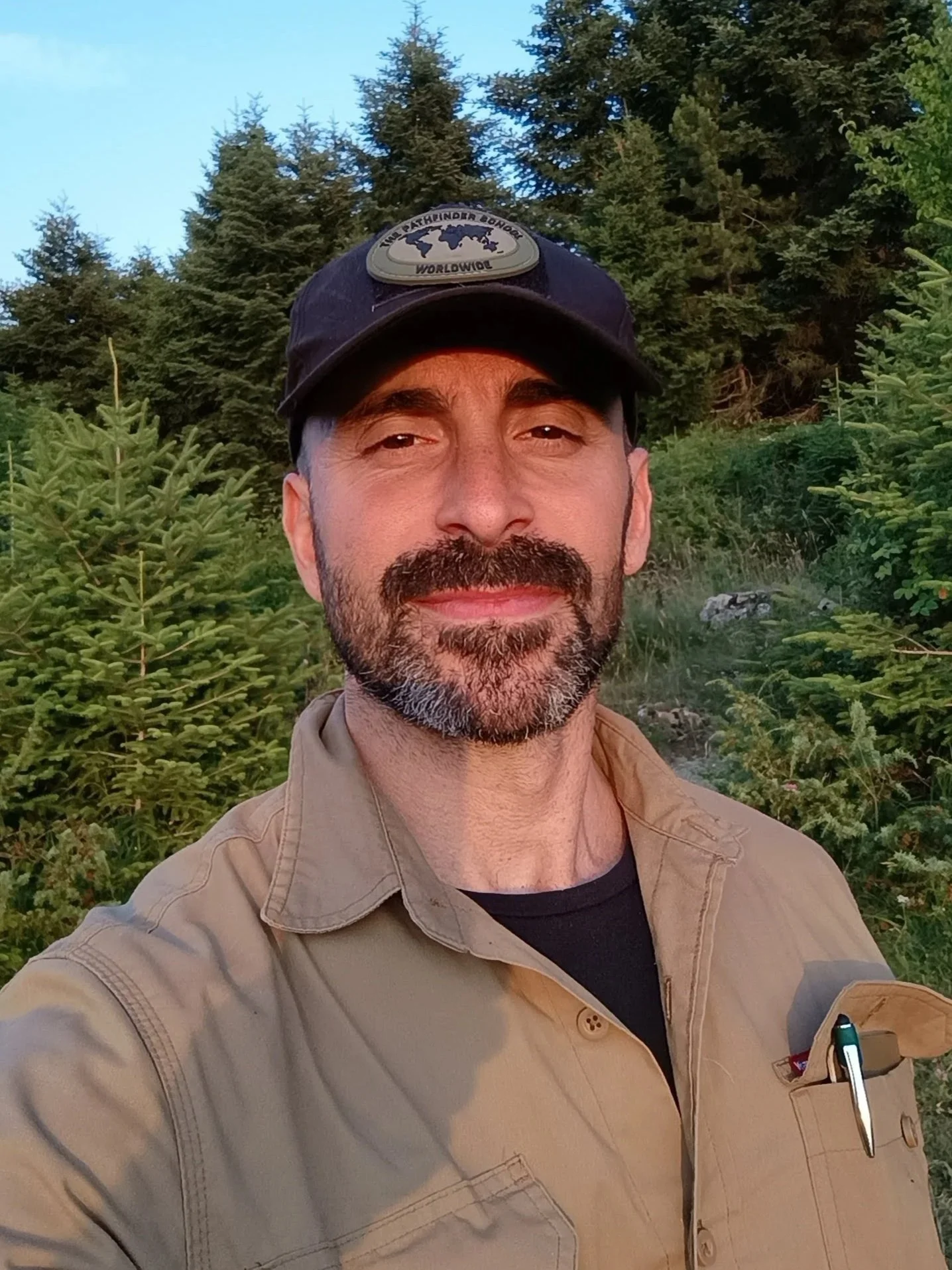 A middle-aged man with a beard and mustache taking a selfie outdoors in a forested area with green trees and clear blue sky in the background. He's wearing a black cap with a badge and a beige shirt with a pocket holding a pen.