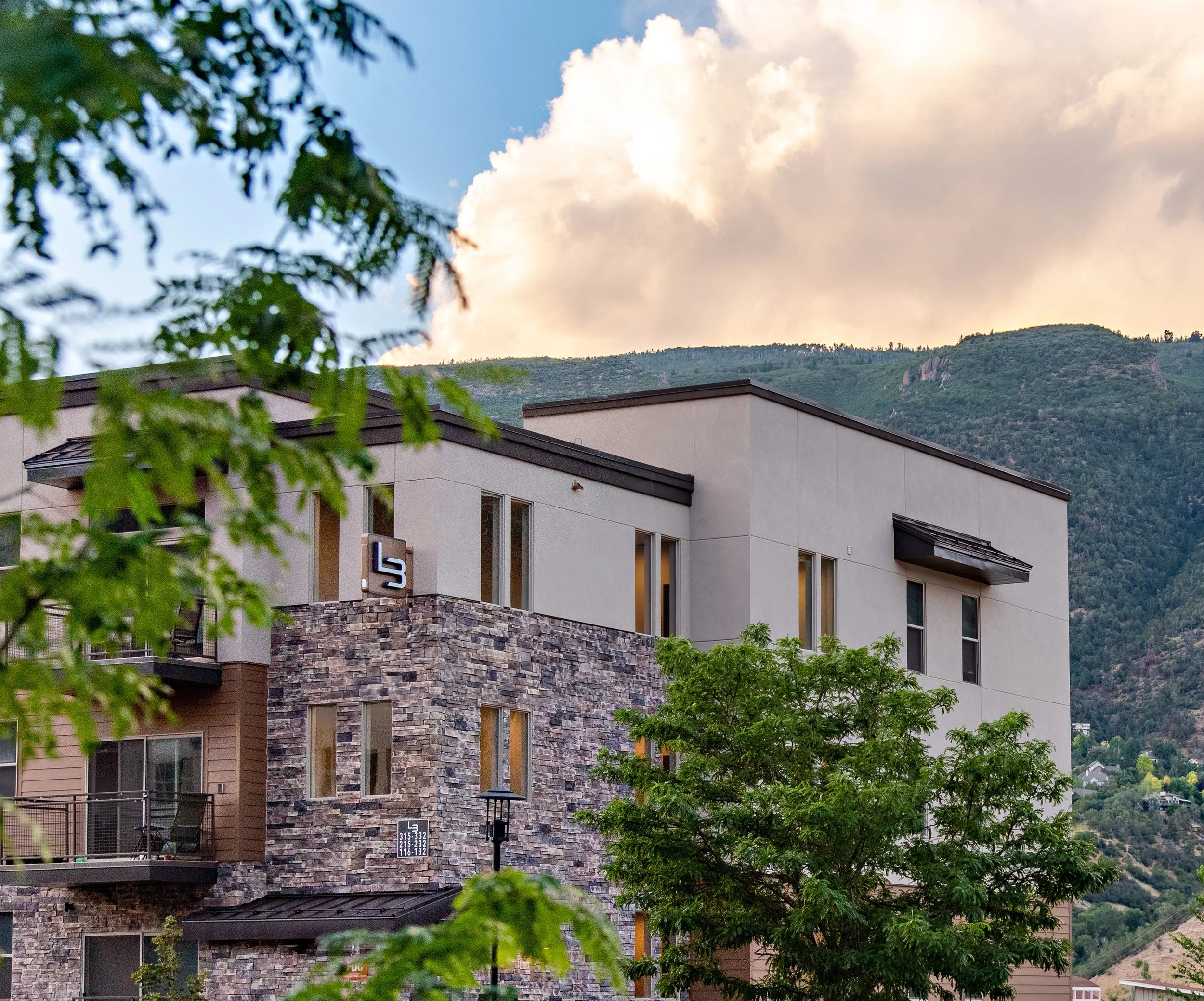 Modern multi-story residential building with stone and beige exterior, large windows, and balconies, set against a backdrop of green mountains and a partly cloudy sky.