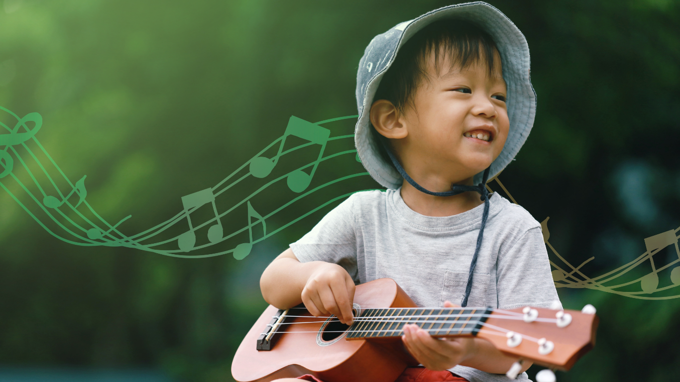Young Asian boy wearing a grey t-shirt and a wide-brimmed hat, smiling while holding a small guitar outdoors with green blurred background and musical notes overlay.