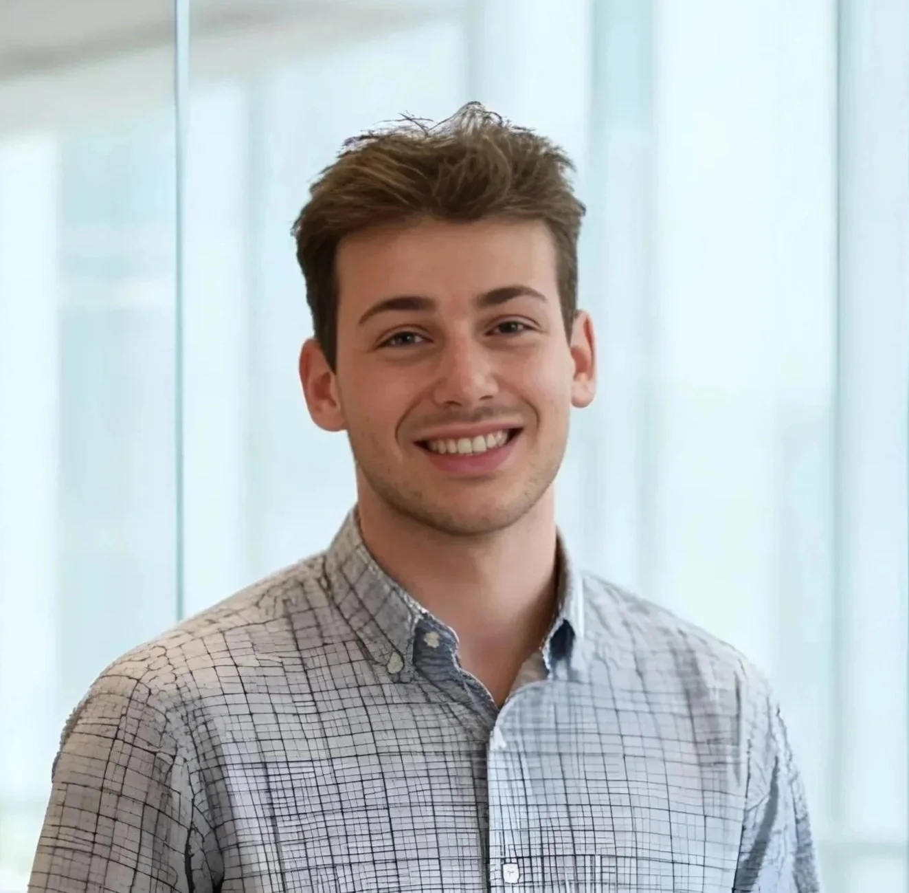 Young man with brown hair smiling in front of frosted glass windows.