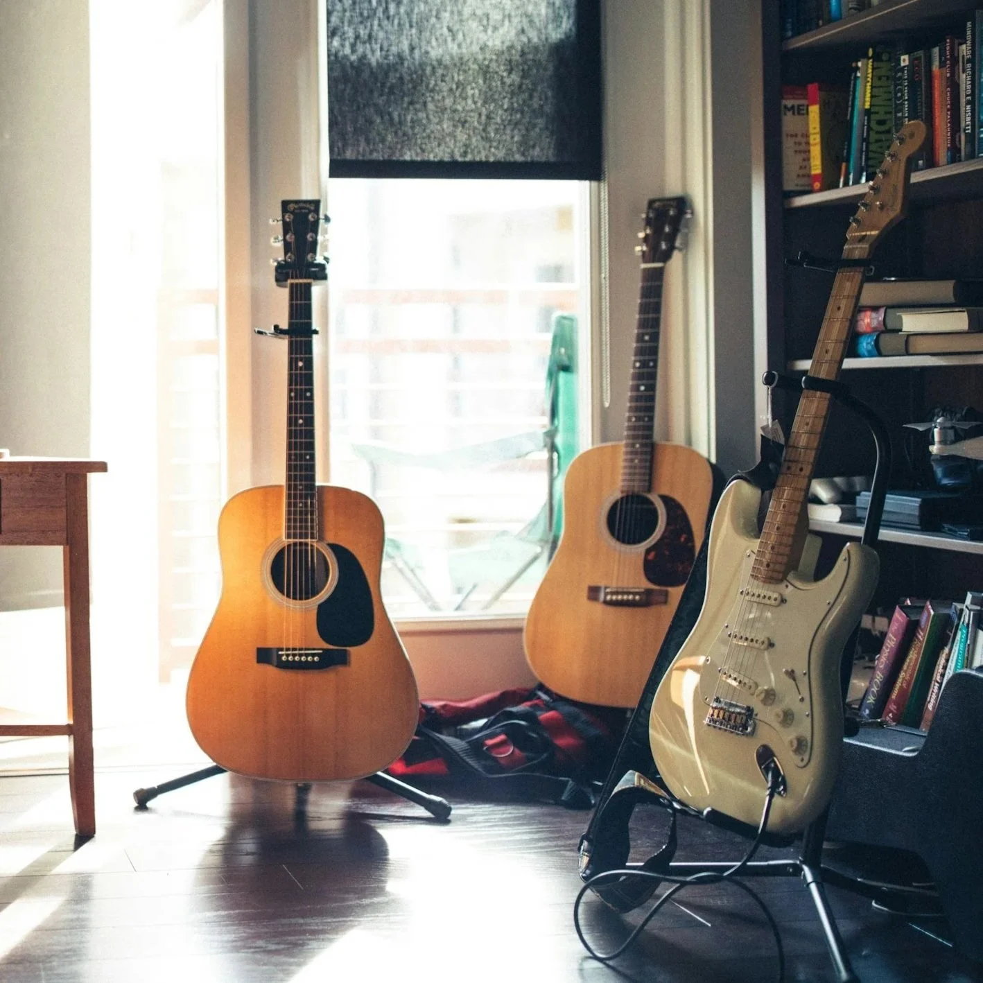 Three guitars in a room near a window. Two acoustic guitars and one electric guitar on a stand. A bookshelf with books and a black electronic device nearby.