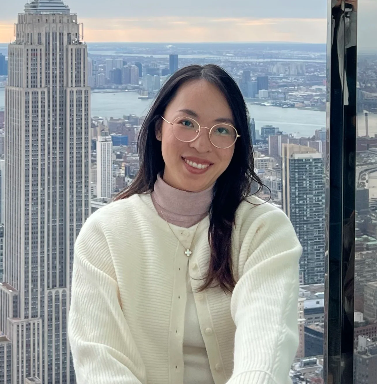 A woman with glasses and a cream-colored cardigan smiling in front of a city skyline with tall buildings and a river in the background.