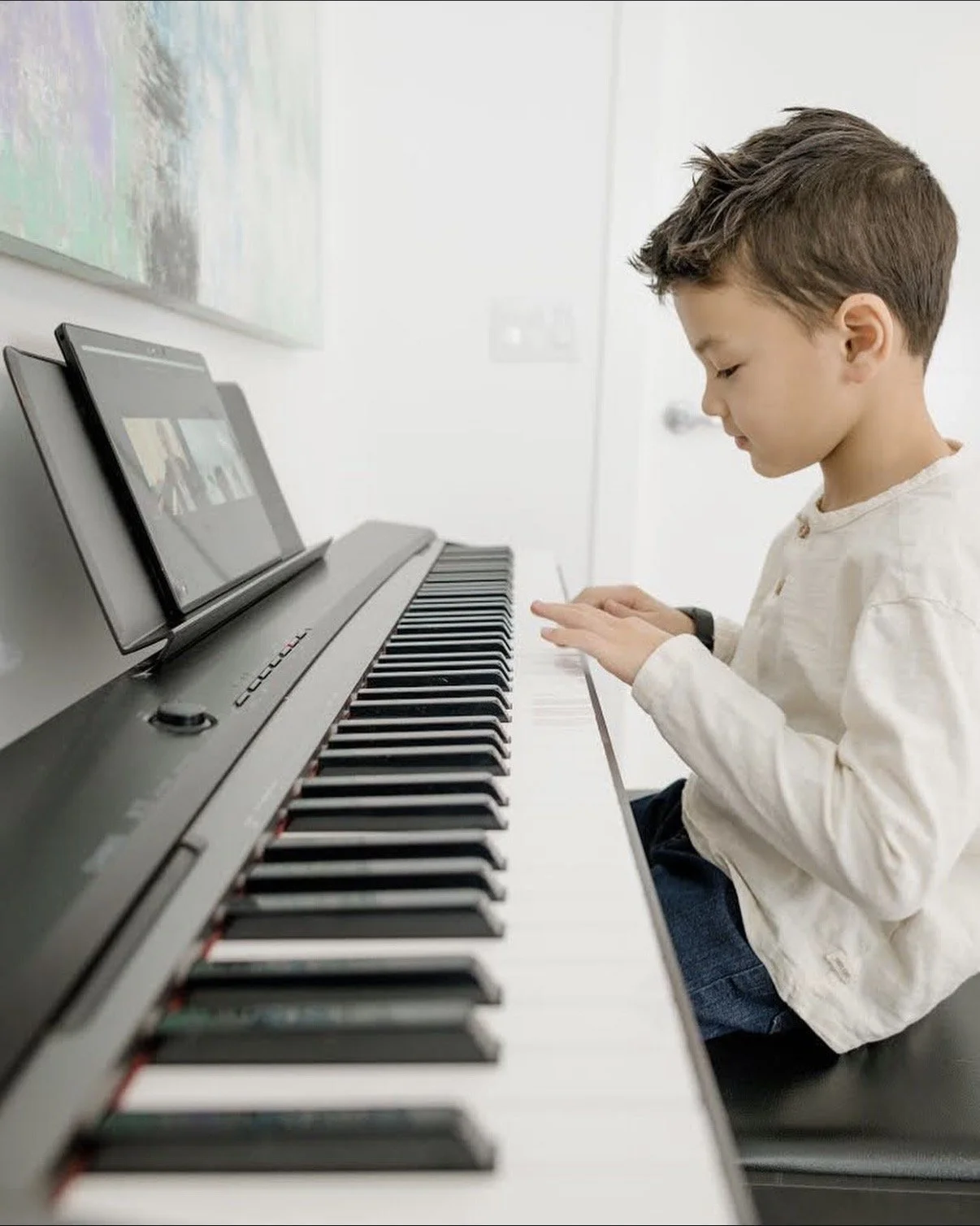 A young boy playing an electronic keyboard with a tablet propped up on it in a bright room.