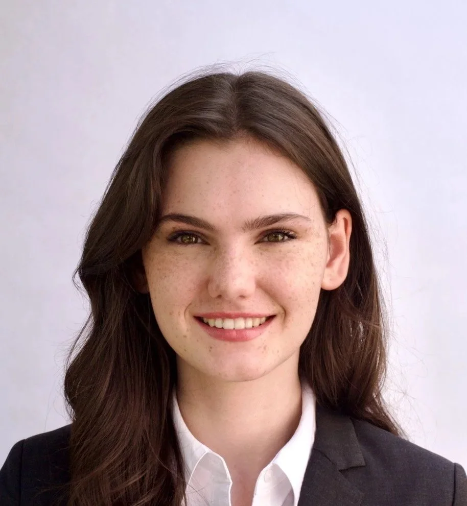 Portrait of a young woman with long brown hair, wearing a black blazer and white shirt, smiling at the camera.