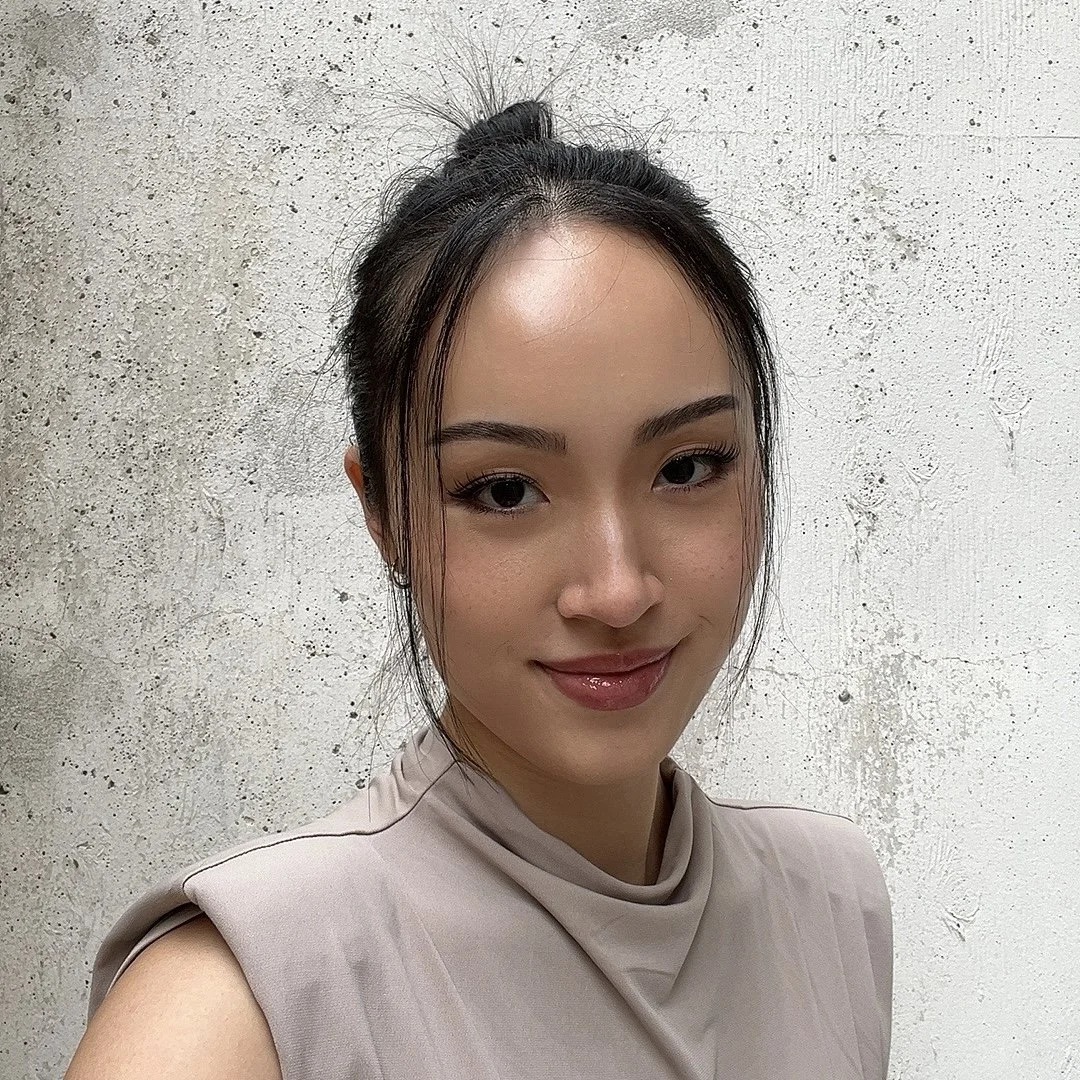 A woman with dark hair tied in a bun, wearing a light beige sleeveless top, smiling and standing against a textured white wall.