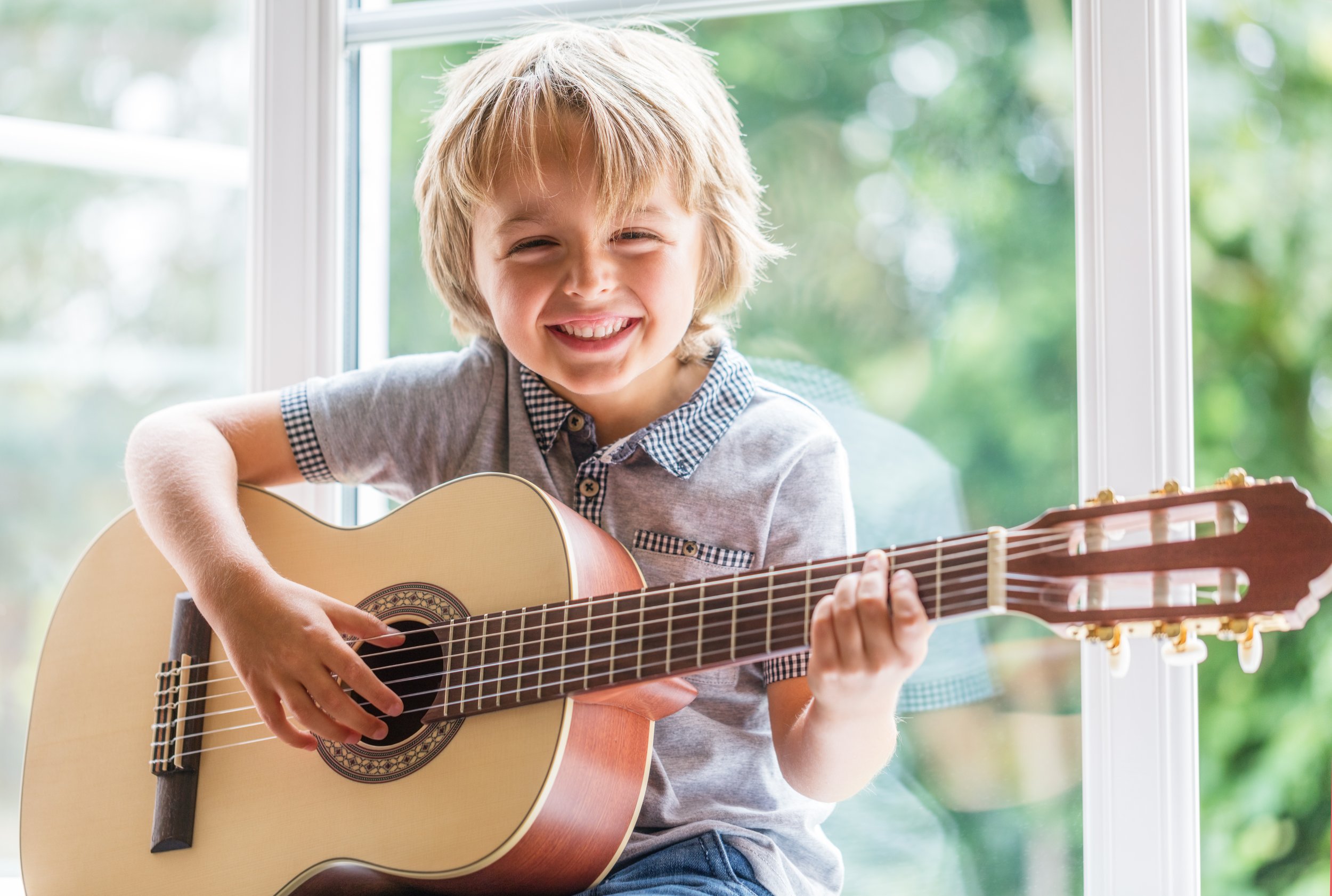 A young boy with blonde hair smiling and playing an acoustic guitar by a window with green trees outside.