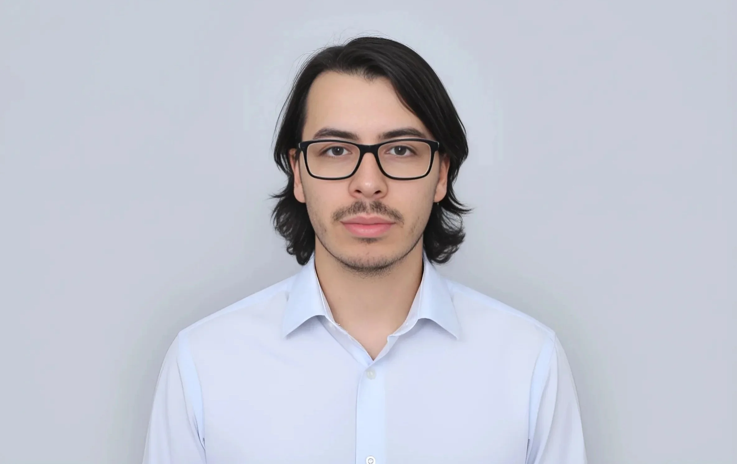 A young man with long dark hair, wearing glasses and a white shirt, standing in front of a plain light gray background.