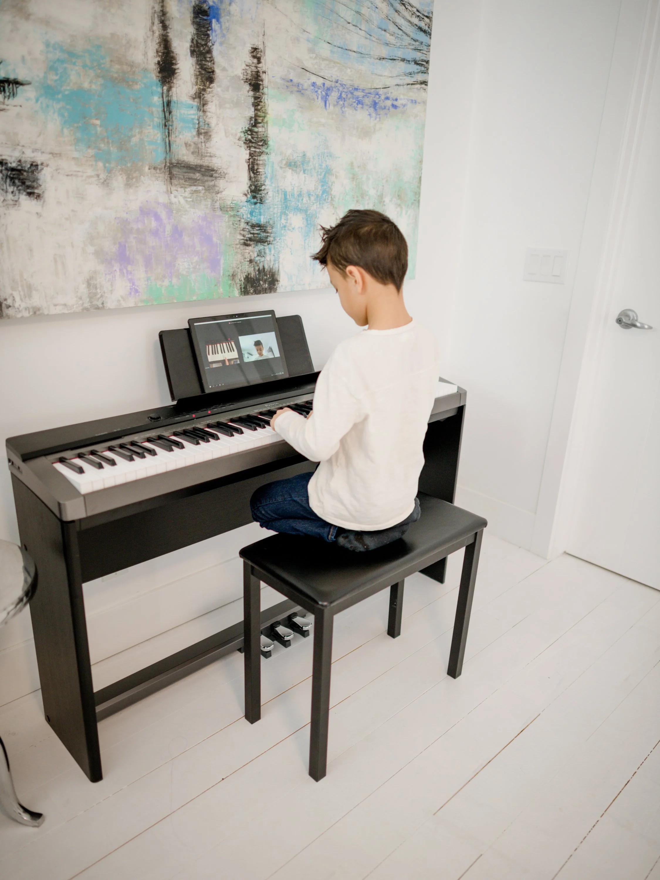 A young boy sitting on a black bench at a digital piano, looking at a tablet displaying a music tutorial. The room has white walls, a colorful abstract painting on the wall, and white flooring.