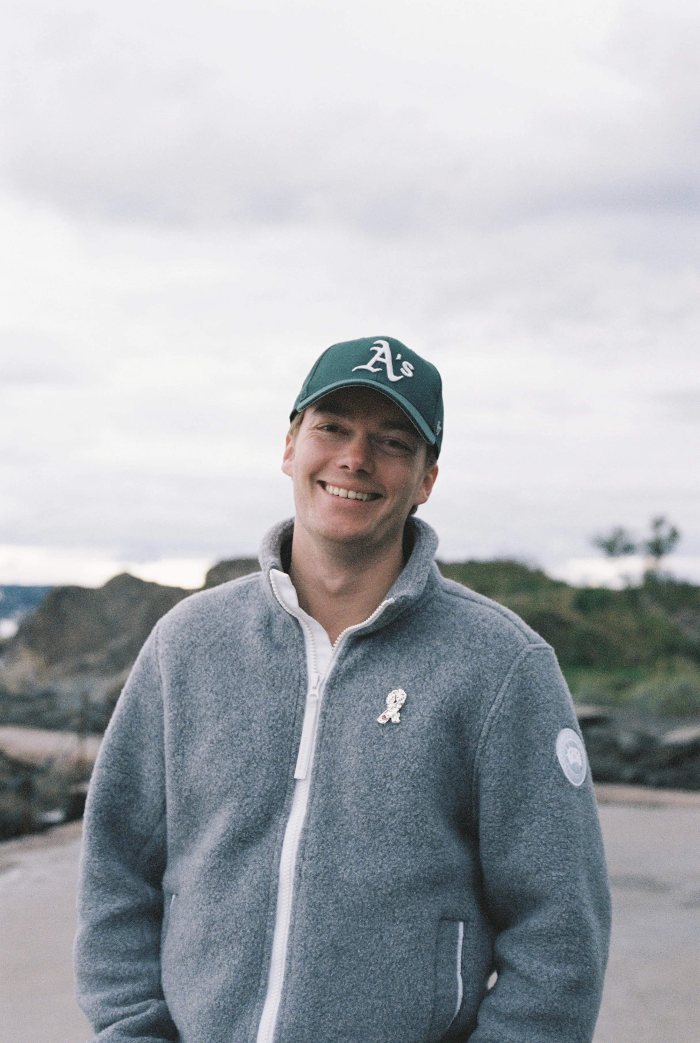 A smiling young man wearing a teal Oakland Athletics baseball cap and a gray zip-up fleece jacket standing outdoors with a rocky landscape and cloudy sky in the background.