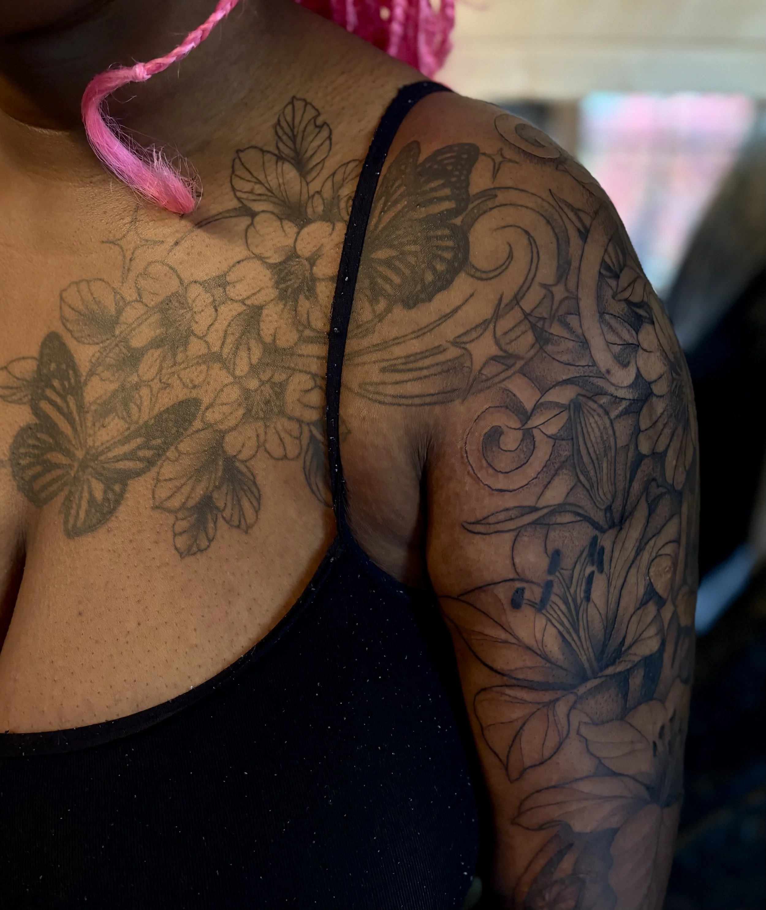 Close-up of a person's shoulder and upper arm showing intricate black floral and butterfly tattoos.