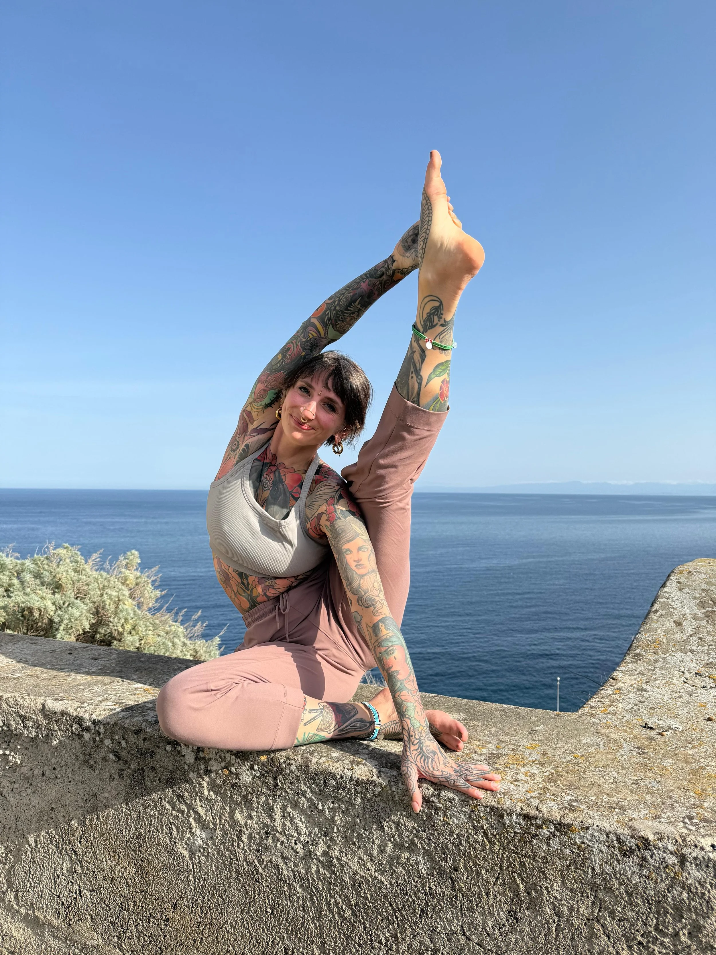 A woman with tattoos practicing yoga outdoors on a stone ledge overlooking the ocean, performing a standing split pose with one leg extended upwards and her head tilted, smiling at the camera.