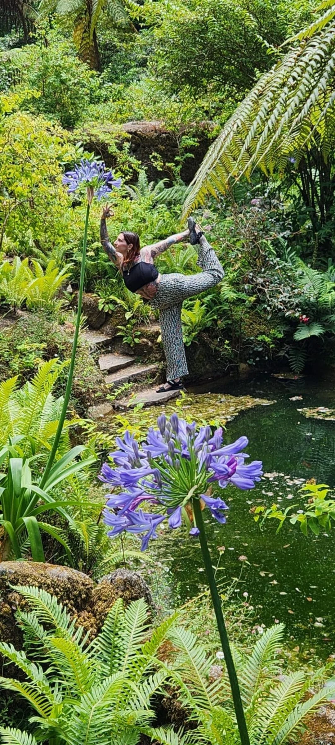 A woman practicing yoga outdoors in a lush garden with tropical plants, purple flowers, and a pond.
