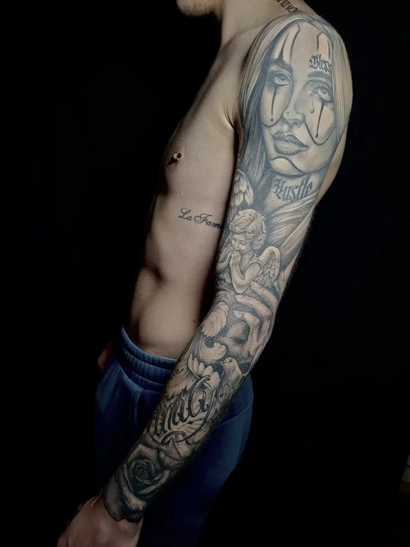 Side view of a shirtless person with tattoos on their arm and chest, standing against a dark background.
