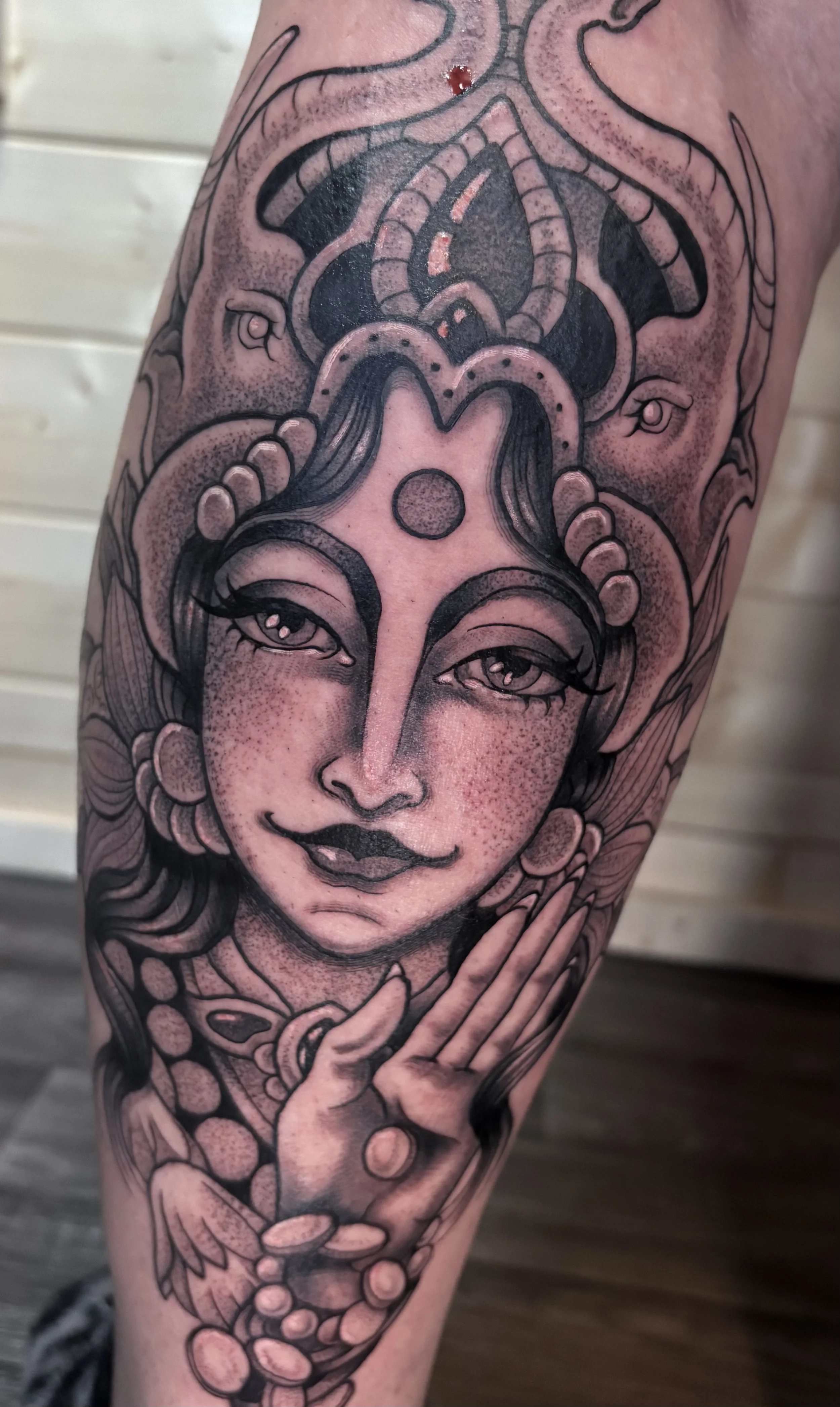 Black and gray tattoo of a serene goddess with hands in prayer position, surrounded by intricate patterns, jewelry, and a decorative headpiece.