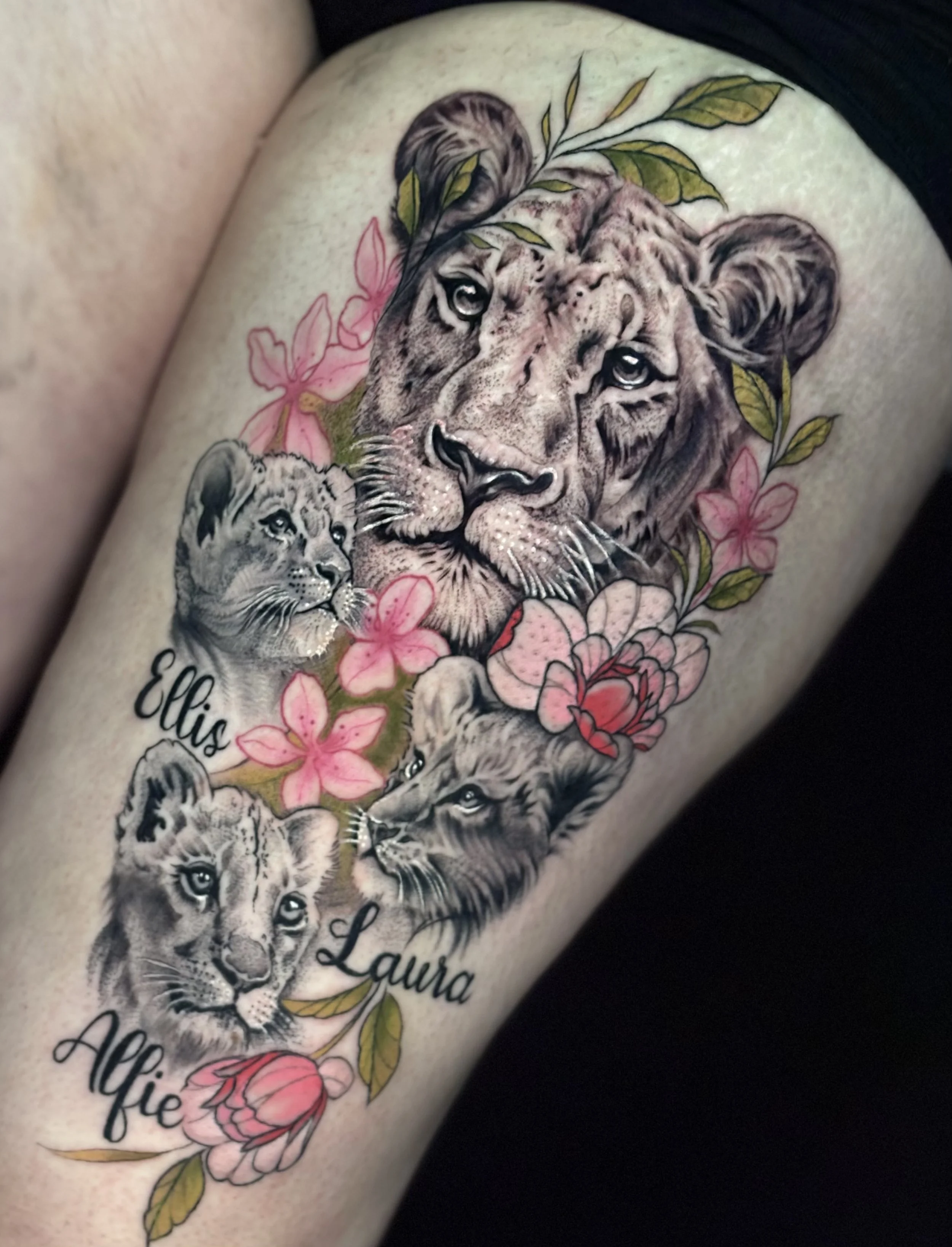 Tattoo of four lion cubs and an adult lion surrounded by pink flowers and green leaves, with the names Ellis, Laura, and Alfie written in cursive.