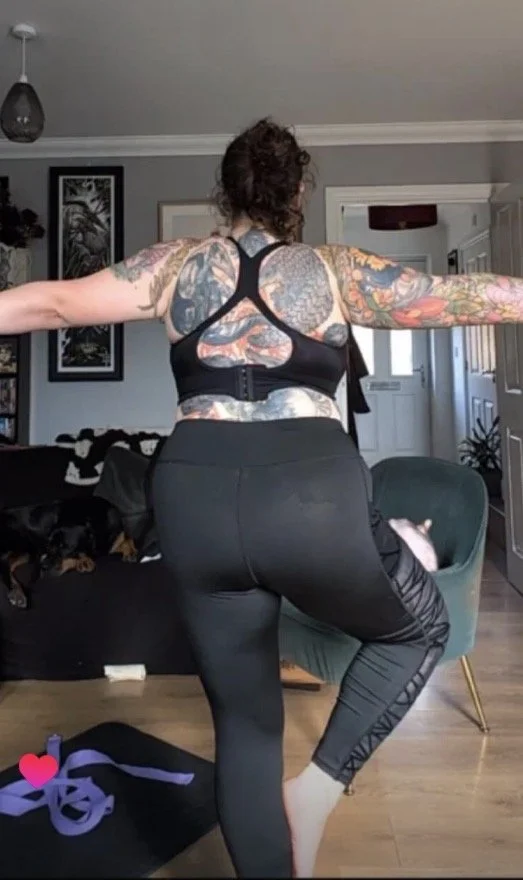 A woman with tattoos stretching her arms and leg in a living room, with a dog resting on a black couch nearby.