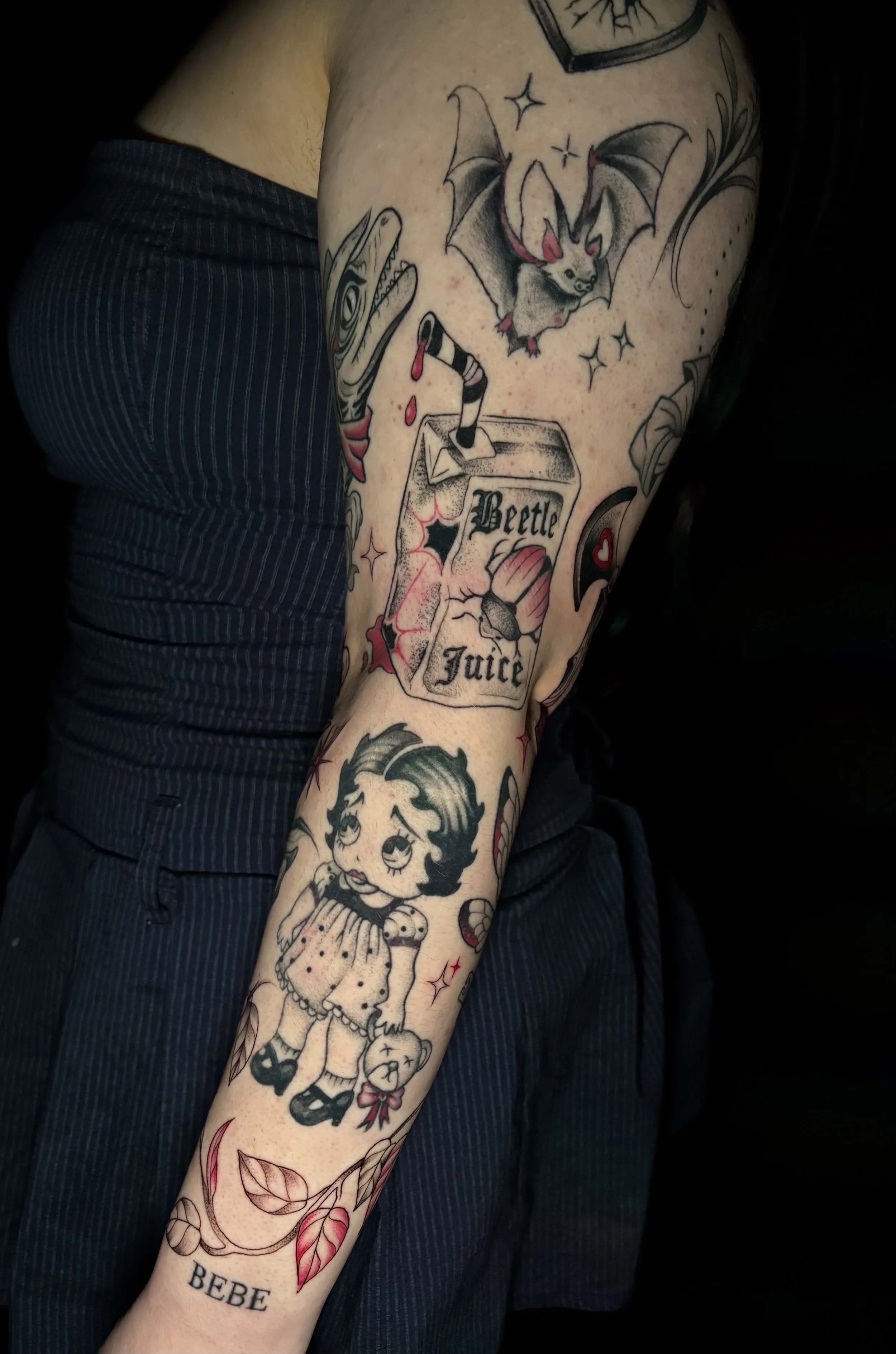 Tattooed arm featuring a cartoon girl holding a teddy bear, a bat, a dinosaur, a juice box labeled 'Beetle Juice', and decorative leaves and stars.