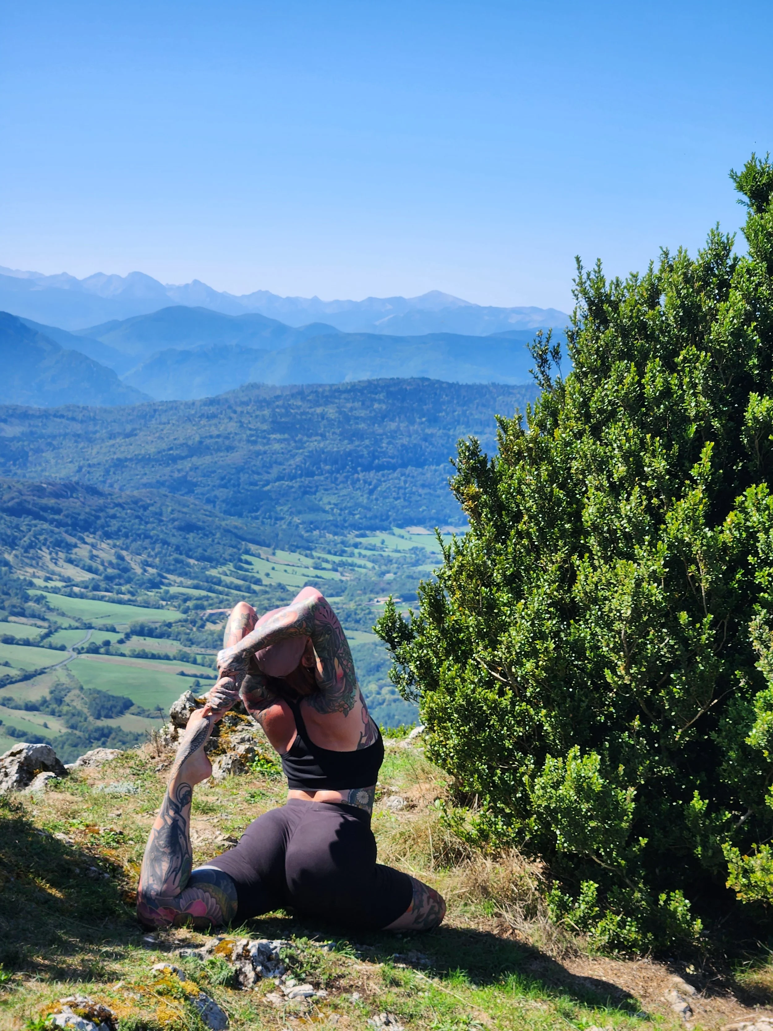 A woman with tattoos practicing yoga outdoors on a mountain, with a scenic view of rolling hills and distant mountains under a clear blue sky.