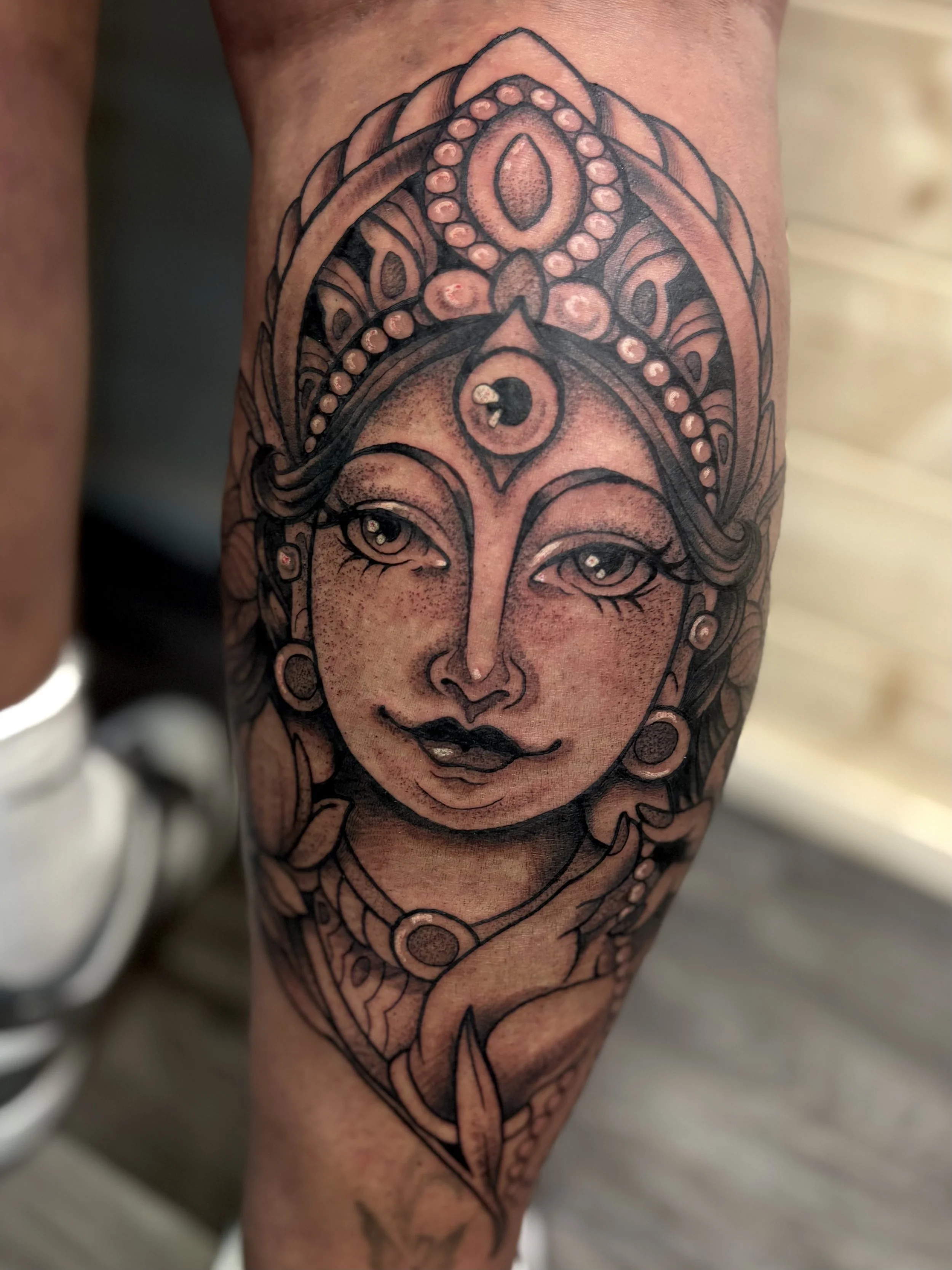 Tattoo of a woman's face with elaborate headpiece, earrings, and necklace, featuring detailed line work and shading.