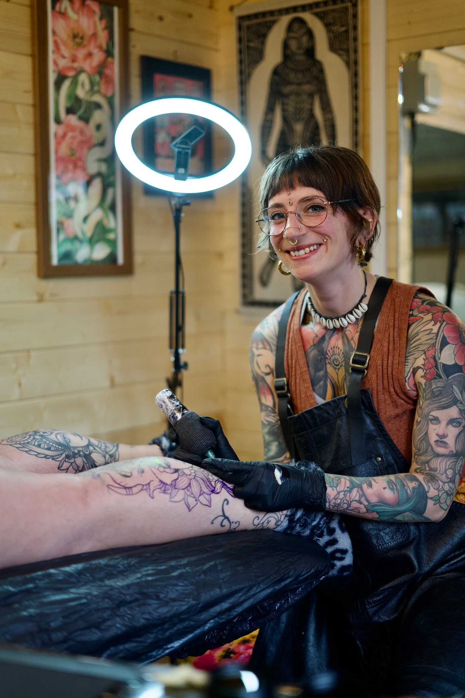 A woman with tattoos and glasses getting a tattoo on her arm in a tattoo studio, smiling at the camera. There is a tattoo machine in her hand, and a ring light is illuminating the scene. The background has wooden walls and framed artwork.