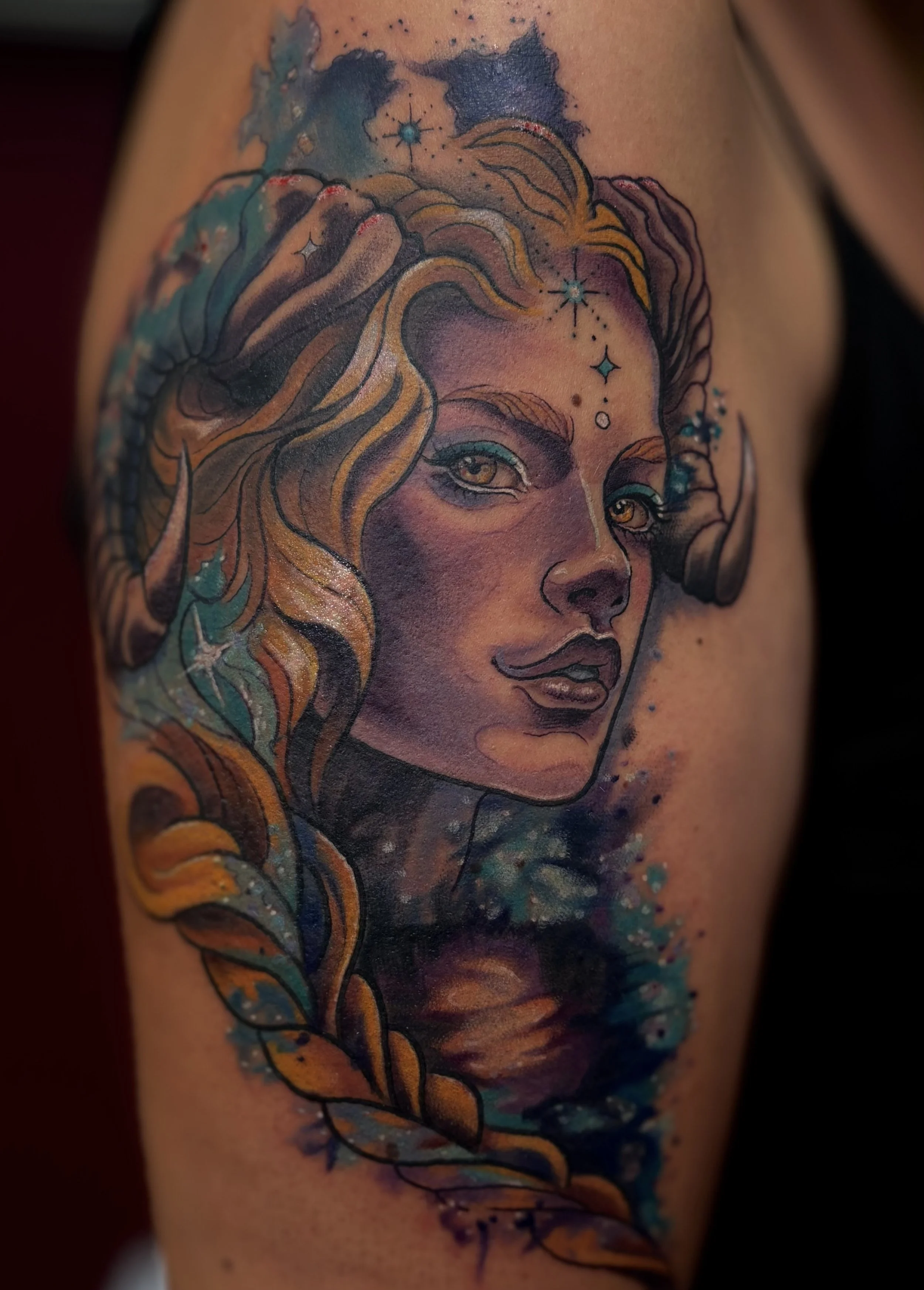 Tattoo of a woman's face with fantasy elements, including horns, colorful hair, and starry celestial designs, on skin.