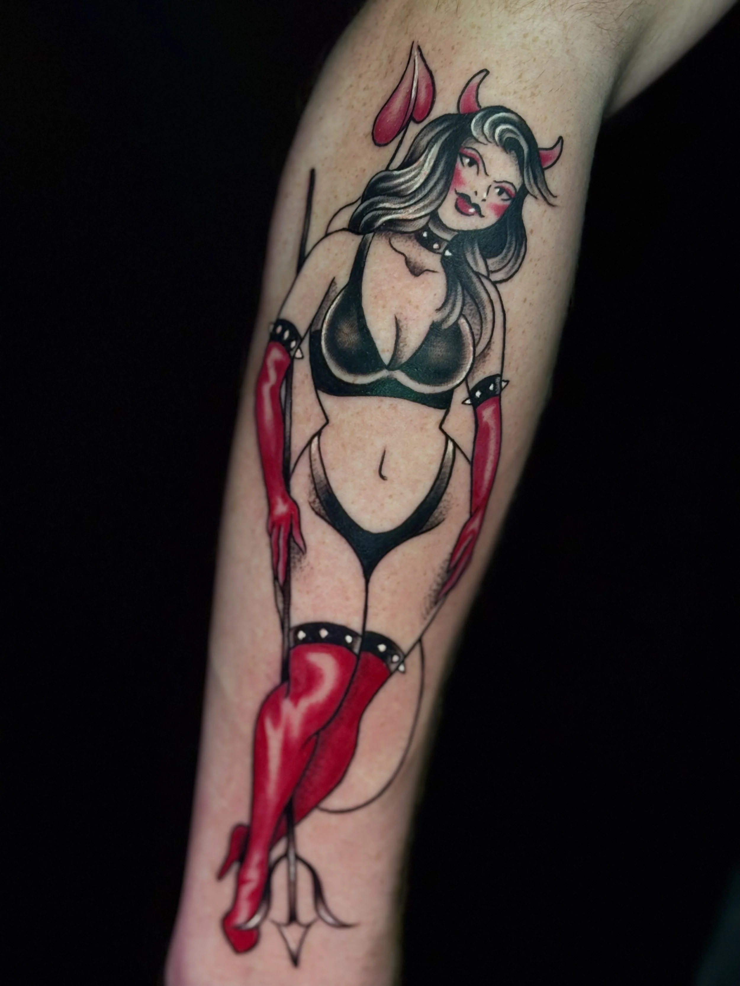 Tattoo of a woman dressed as a devil with pink devil horns, black and gray hair, a black revealing outfit, red gloves, and red thigh-high boots.