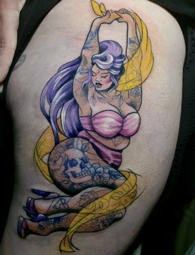 Tattoo of a woman with purple hair, pink top, and tattoos on her arms, sitting with legs crossed, holding a yellow ribbon, with a skull tattoo on her thigh.