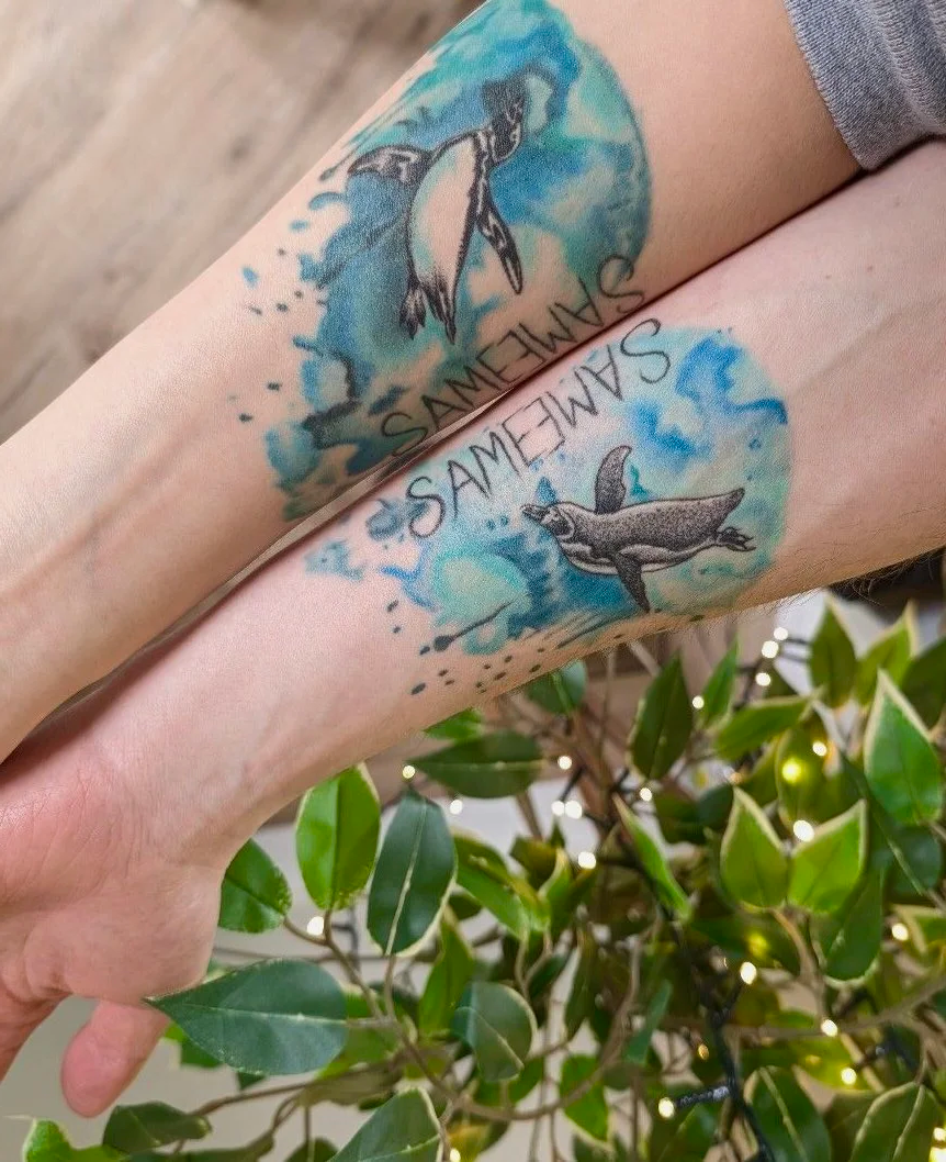 Two arms with matching tattoos of penguins and the word 'SAME' in black ink on a blue watercolor background.