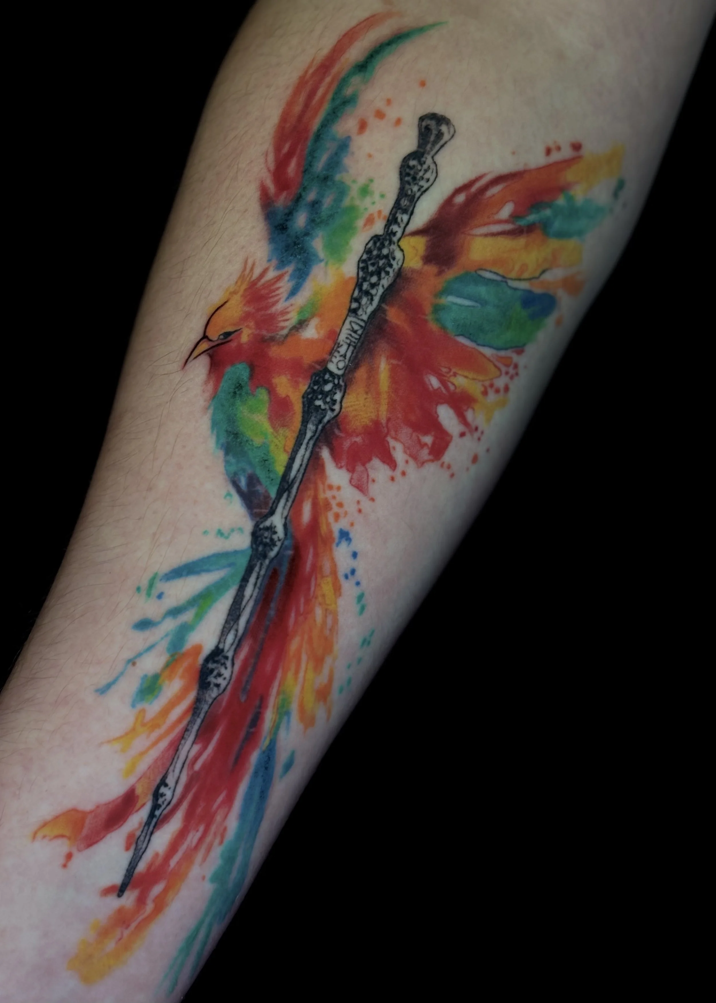 Colorful watercolor tattoo of a phoenix with a black knife blade running through it on a person's arm.