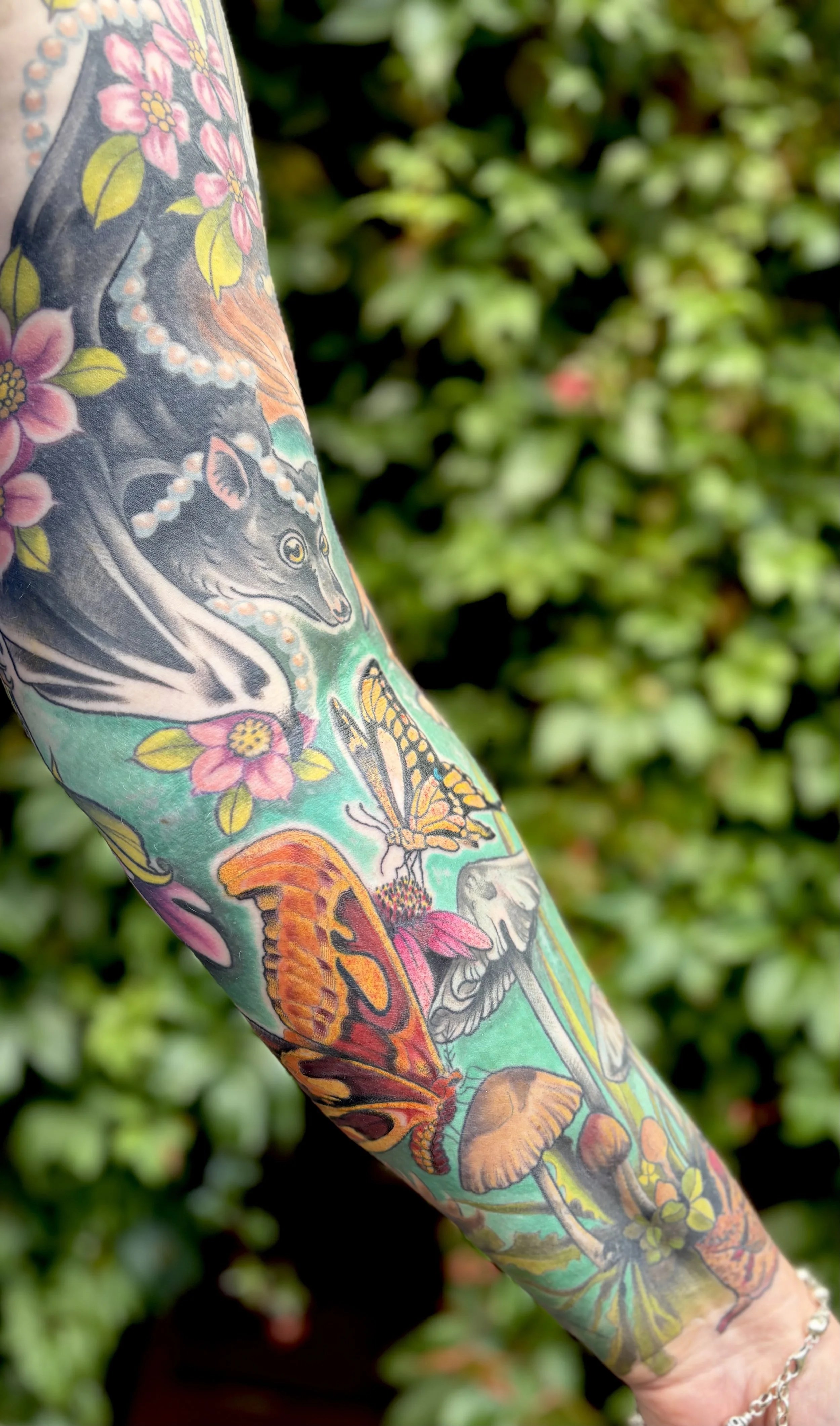 Close-up of a colorful tattoo sleeve featuring a wolf's face, pink flowers, orange and yellow butterflies, and mushrooms, with greenery in the background.