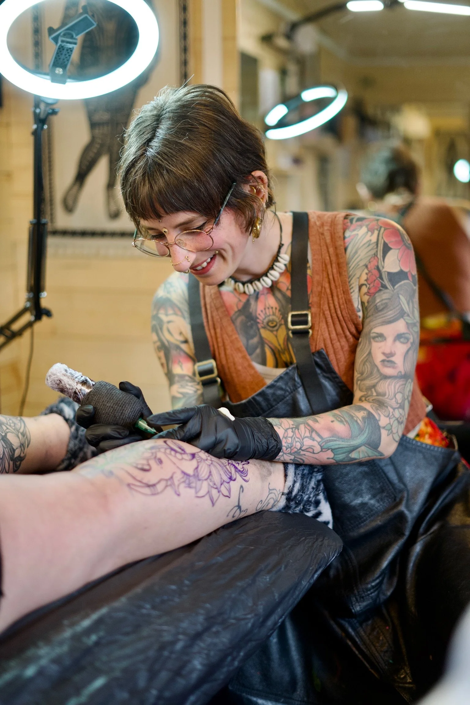 A woman with short brown hair, glasses, and tattoos on her arms is tattooing someone’s leg. She is smiling and wearing black gloves, a shell necklace, and a brown sleeveless top. There are bright ring lights around her, and the setting appears to be a tattoo studio with wooden decor.