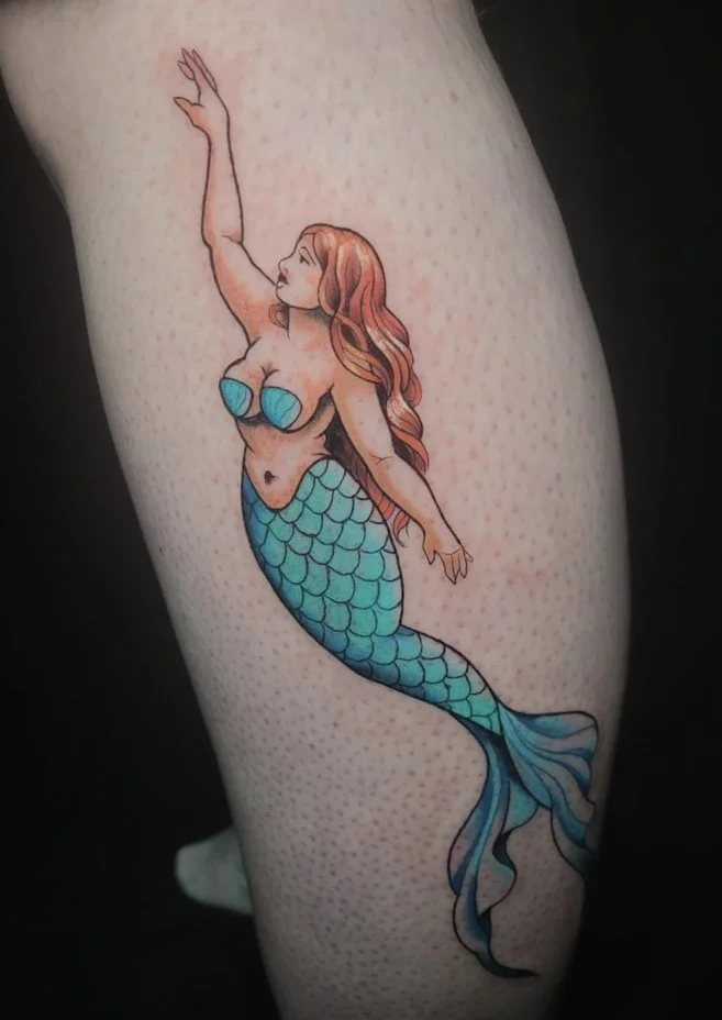 Tattoo of a mermaid with long, wavy red hair, blue seashell top, and blue-green tail, reaching upward with one arm.