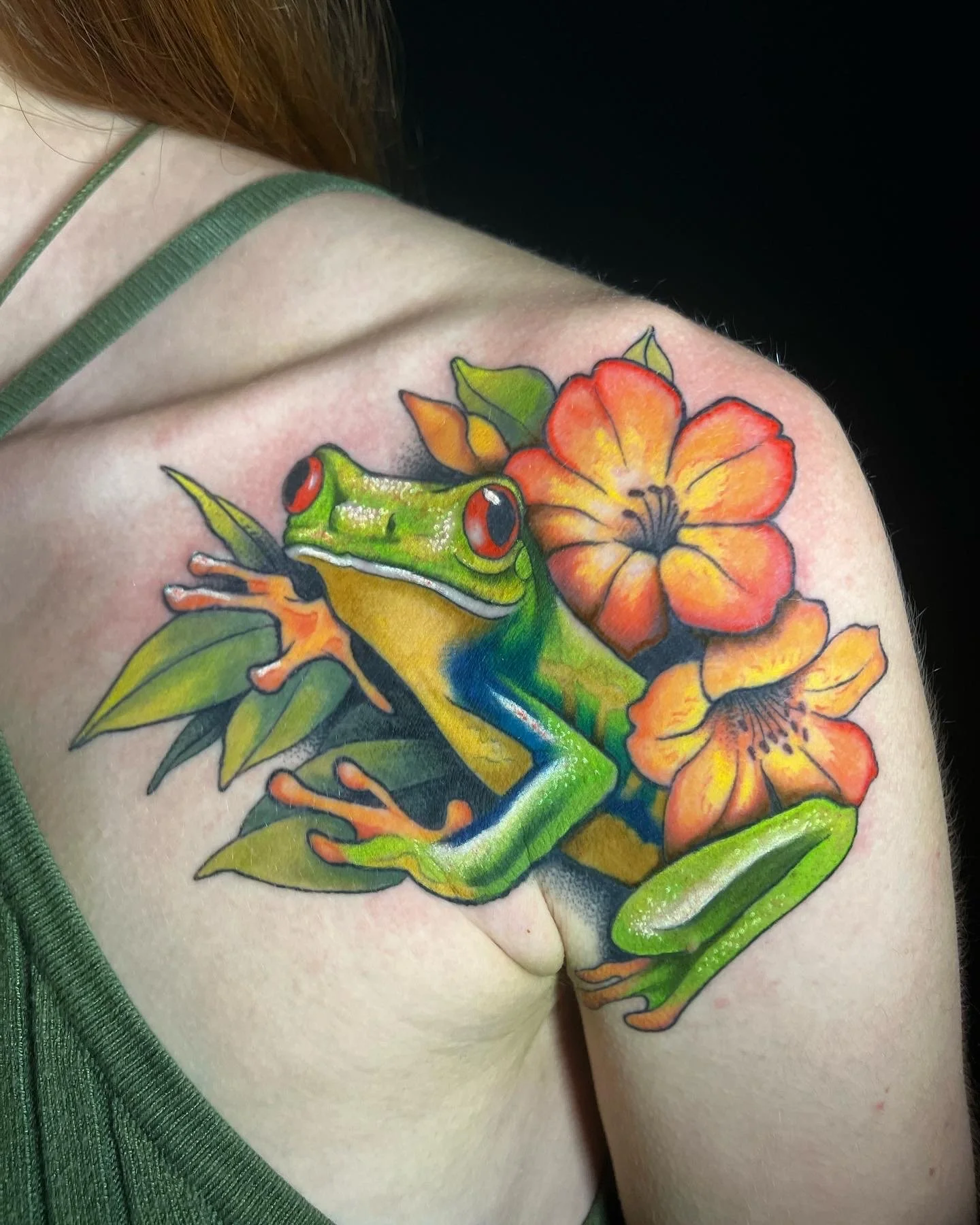 Colorful tattoo of a green frog and orange flowers on a person's shoulder.