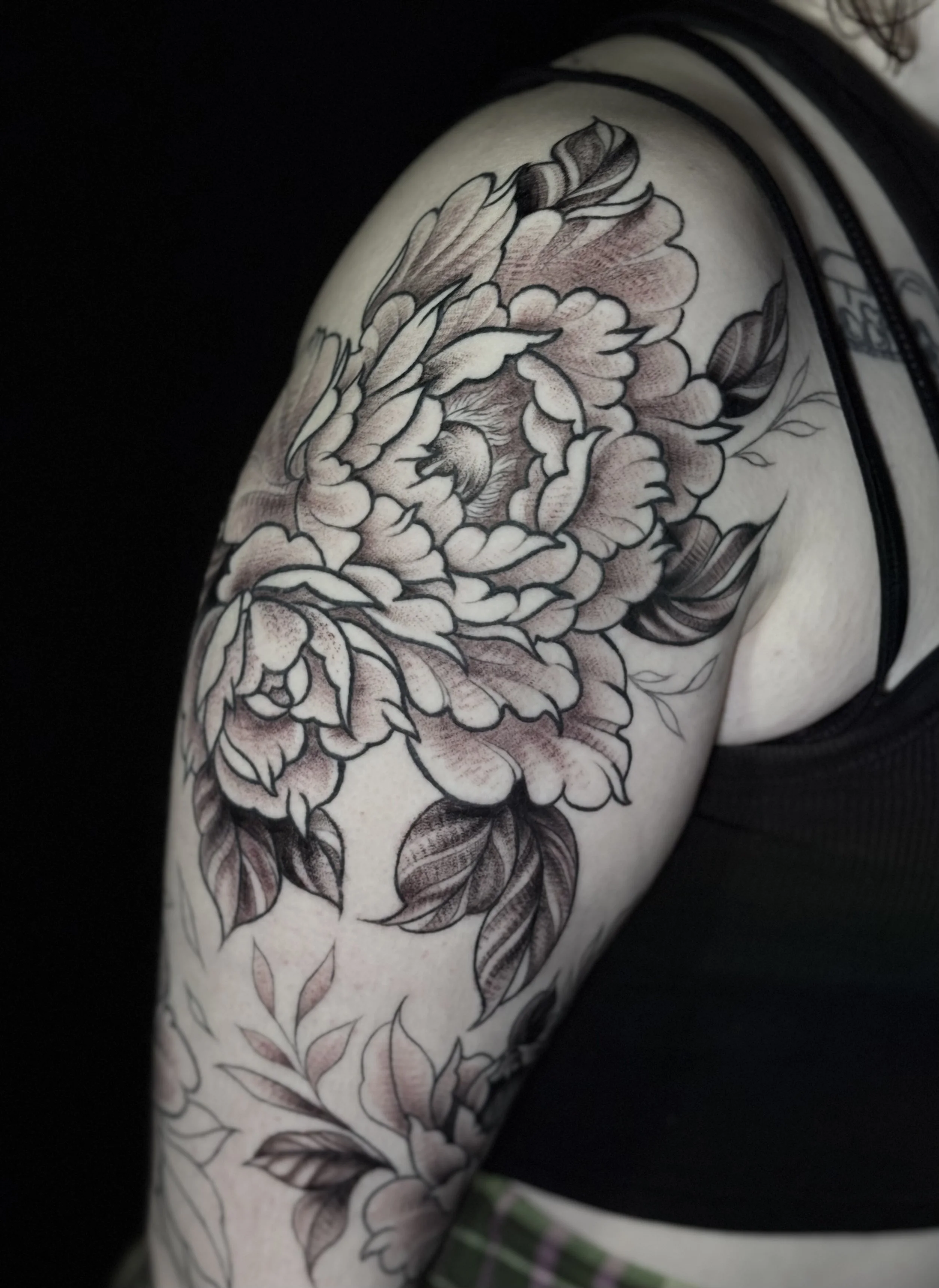 Close-up of a person's shoulder with a large black and gray floral tattoo featuring peonies and leaves.