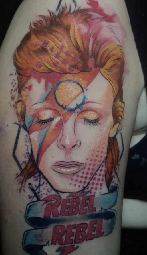 Colorful tattoo of a woman with eyes closed, featuring a lightning bolt on her face, the words 'Rebel Rebel' at the bottom, and a circular design on her forehead.