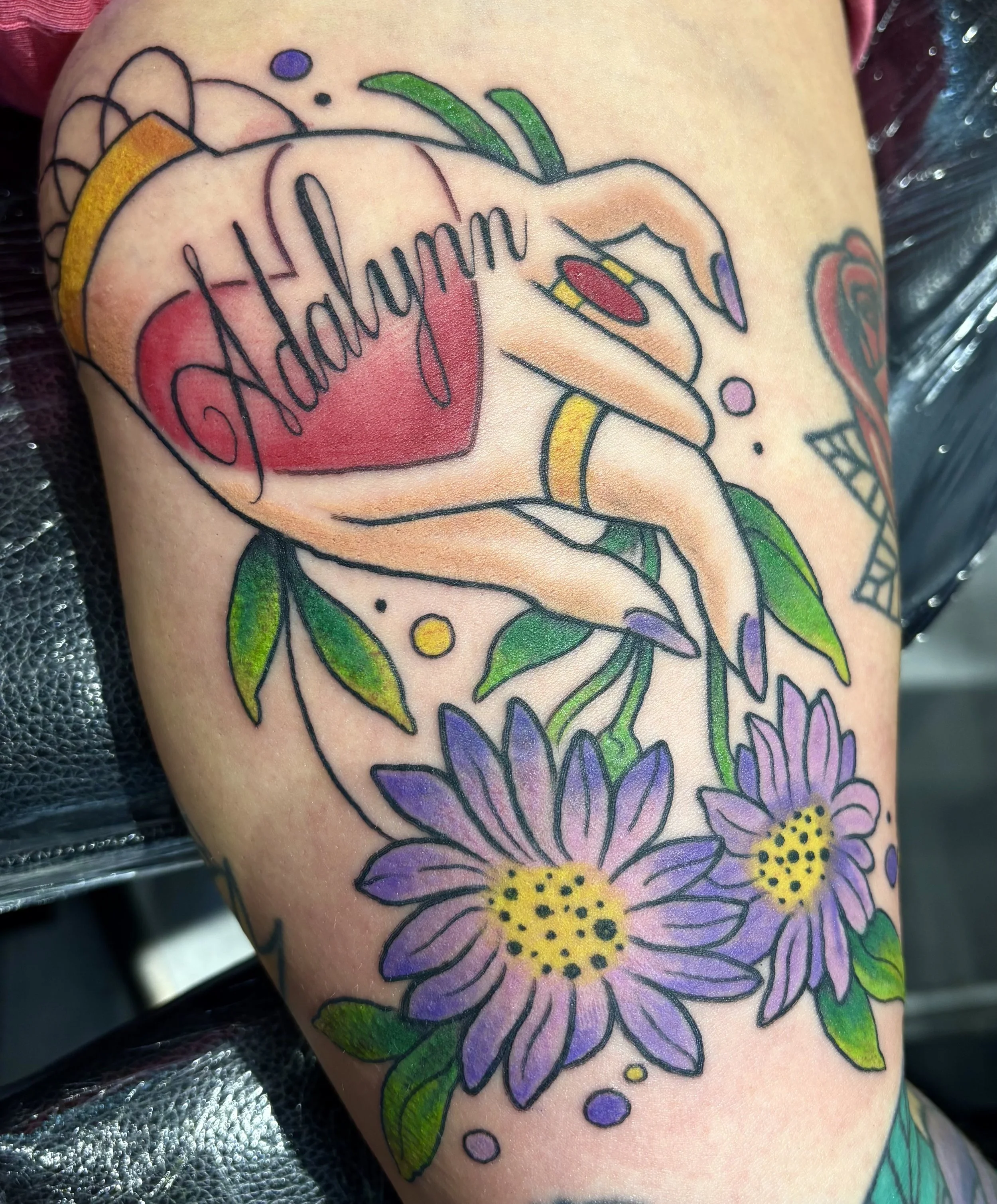 Tattoo of a hand holding a heart with the word 'Addyhum' written inside, surrounded by colorful flowers, leaves, and decorative elements.