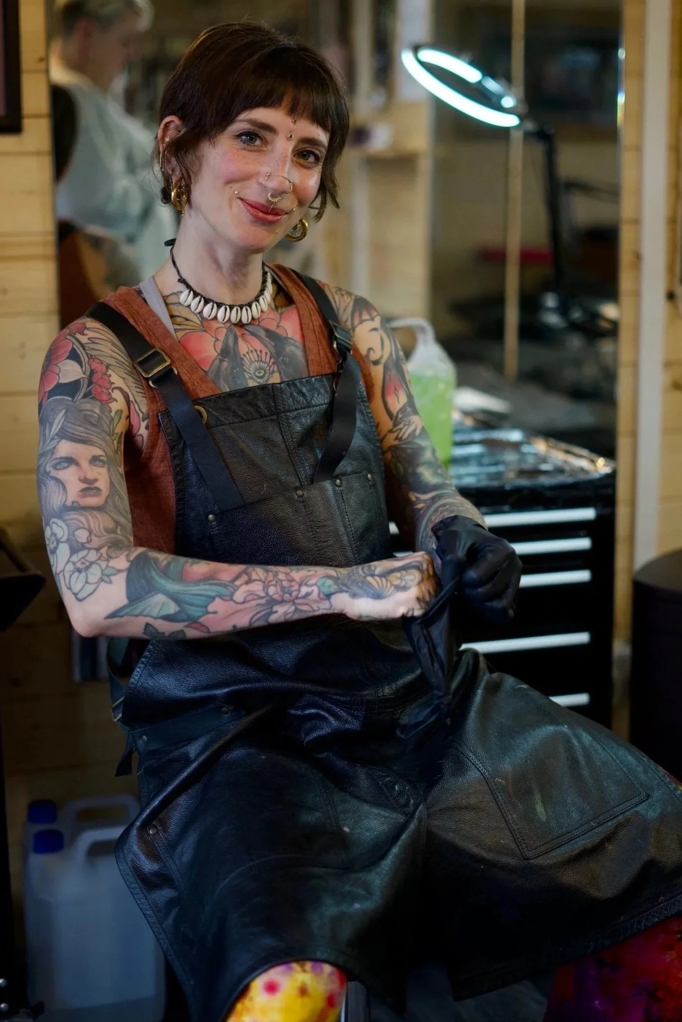 A tattooed woman with short brown hair, smiling, wearing a shell necklace, black gloves, and black leather apron, sitting in a tattoo studio.