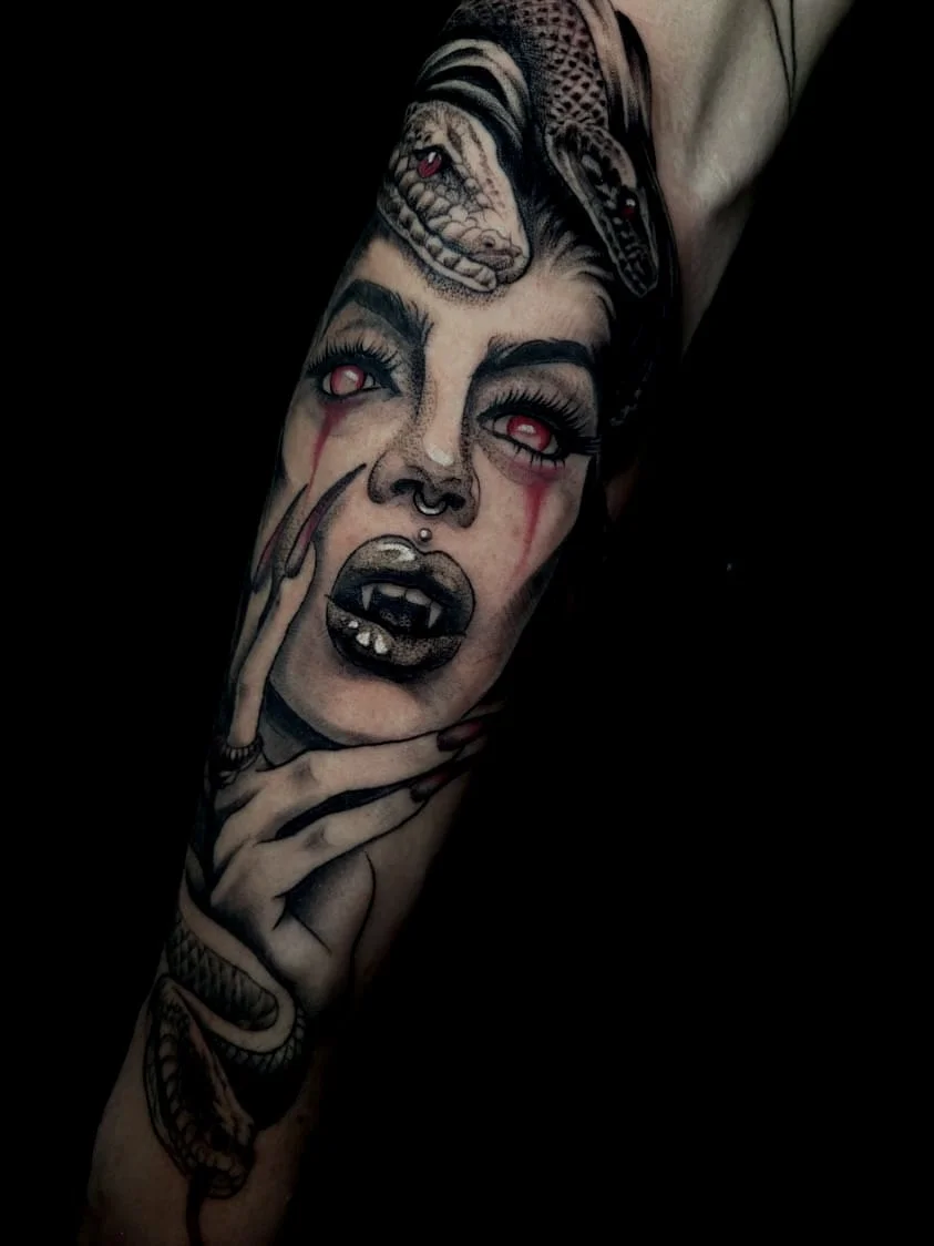 Tattoo of a woman's face with red eyes, blood tears, and open mouth with sharp teeth, holding her chin with a skeletal hand, with a snake coiled around her wrist and a snake head on her forehead.