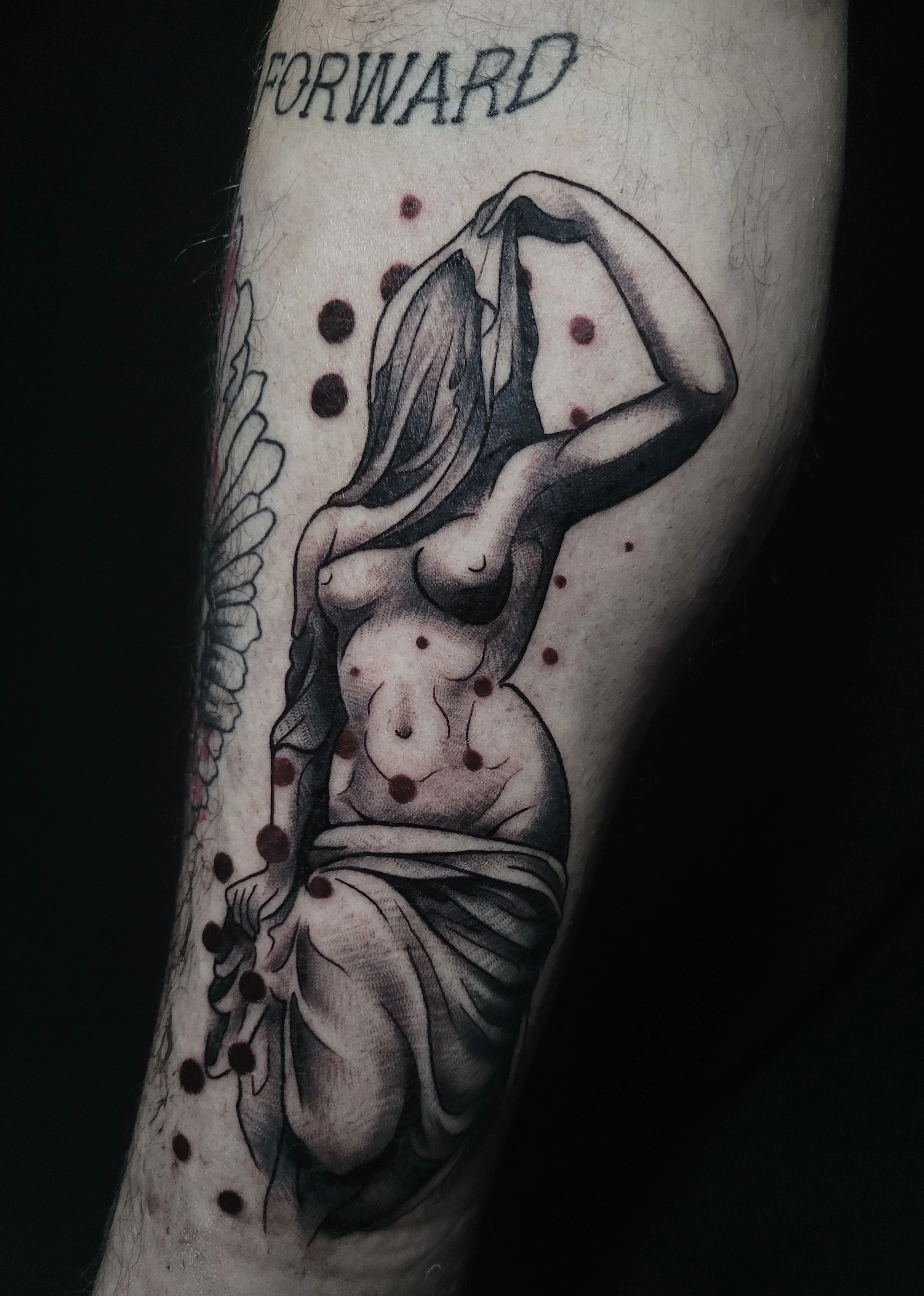 A black and gray tattoo of a woman with wings, sitting with her knees drawn to her chest, with the word "FORWARD" above her head, and red dots scattered around the tattoo.