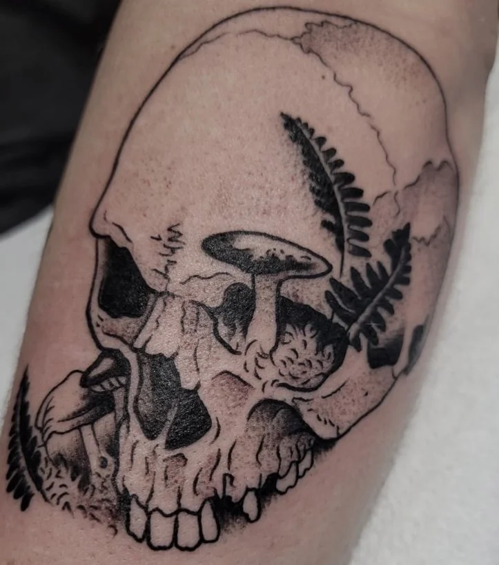 Tattoo of a human skull with mushrooms and fern leaves depicted on it.
