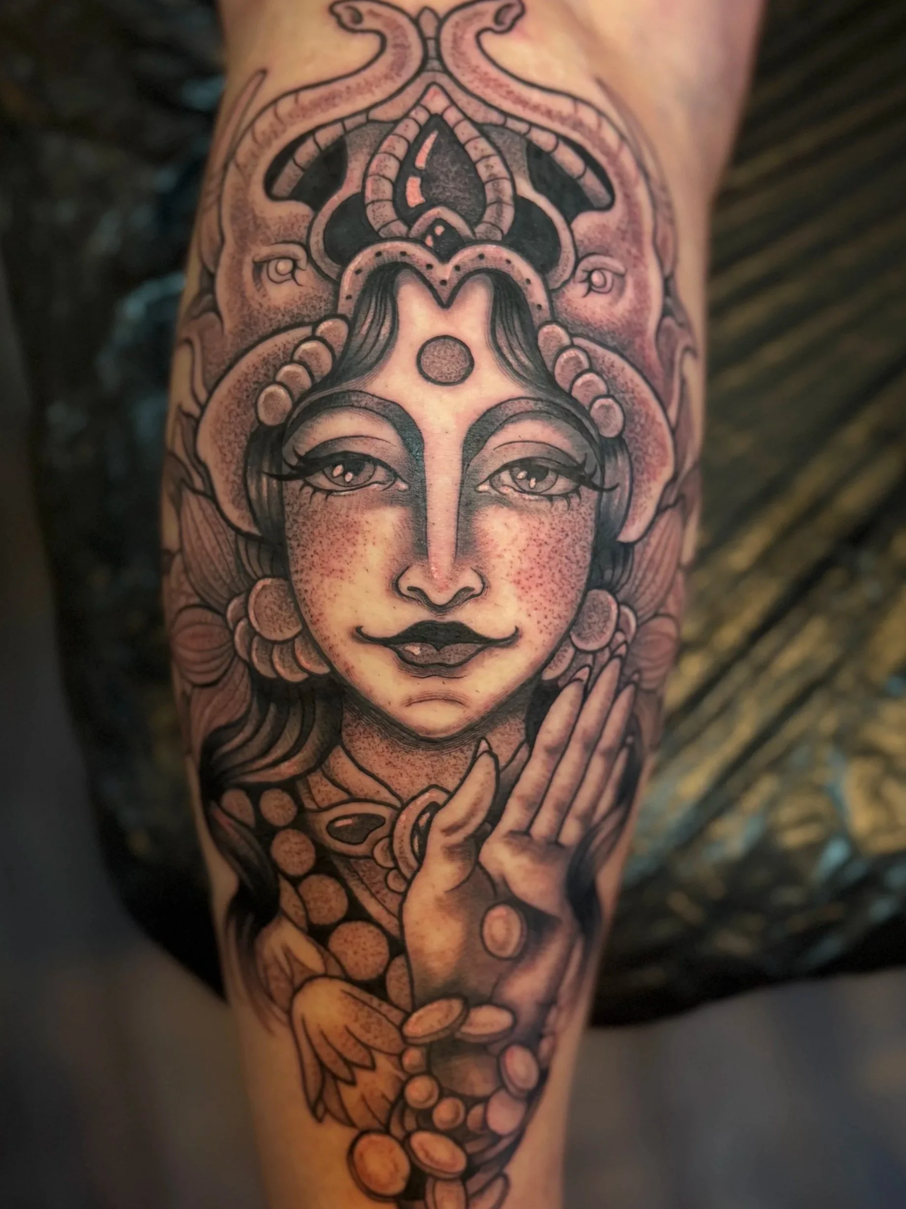 Black and gray tattoo of a woman with detailed facial features, long hair, a third eye on her forehead, and surrounded by ornate decorative elements, including coins and a hand gesture.