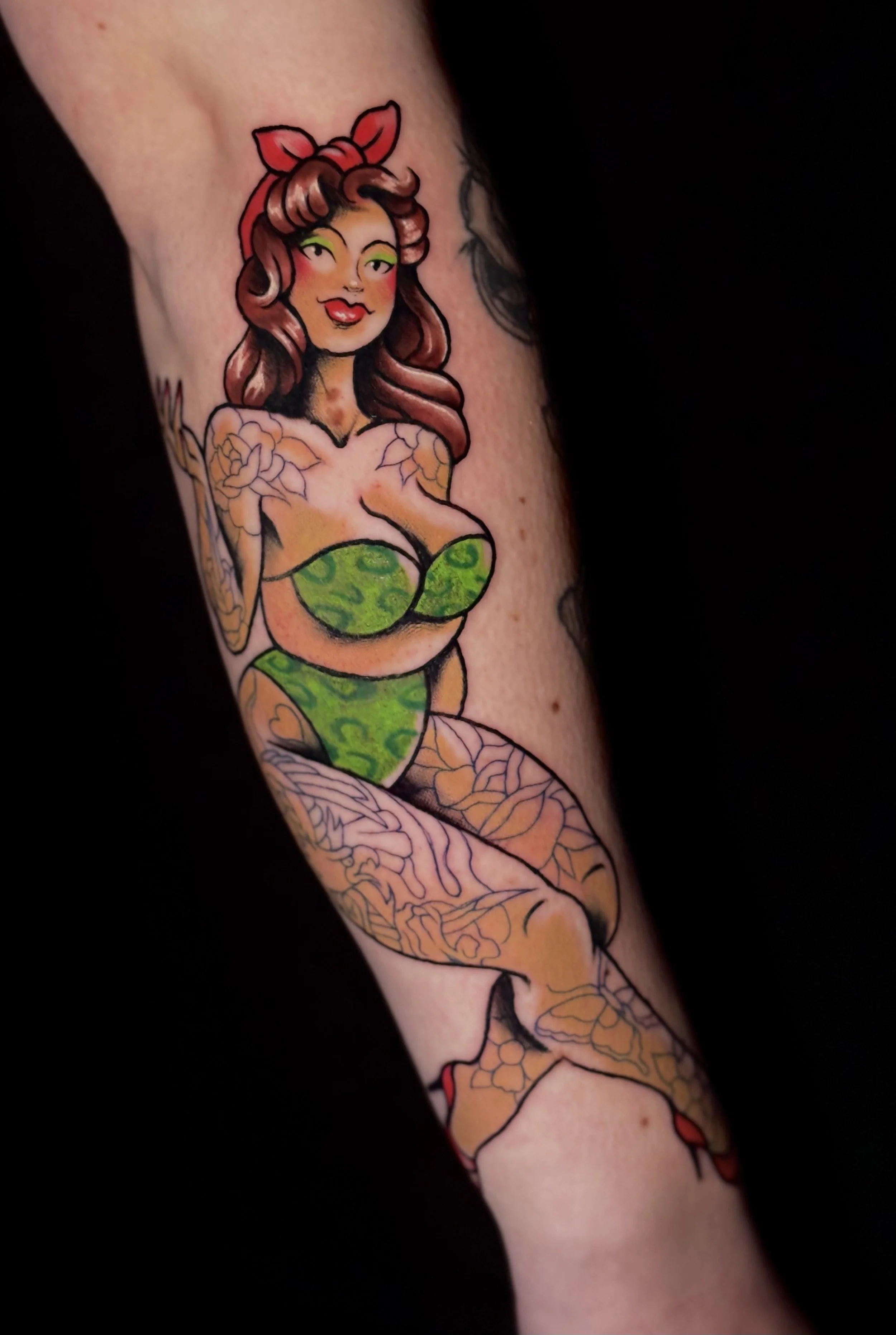 Color tattoo of a pin-up style woman with brown hair, green eyes, red lips, wearing a green leopard print bikini, red headband, and red shoes.