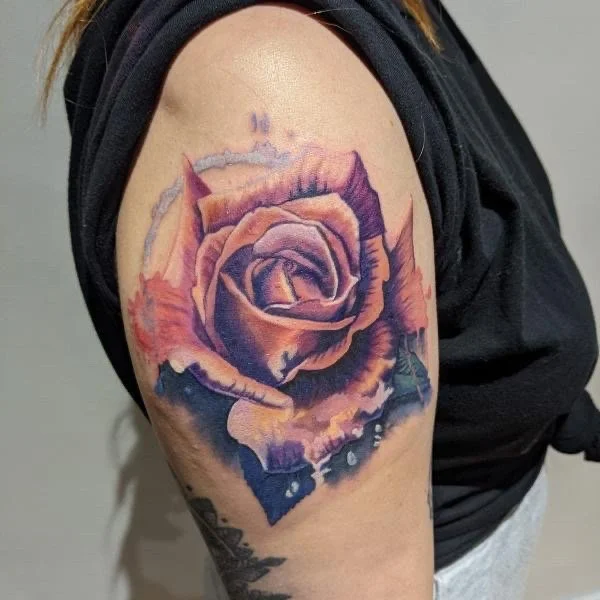 A colorful tattoo of a rose on a person's upper arm, featuring shades of purple, pink, and orange with detailed petals.