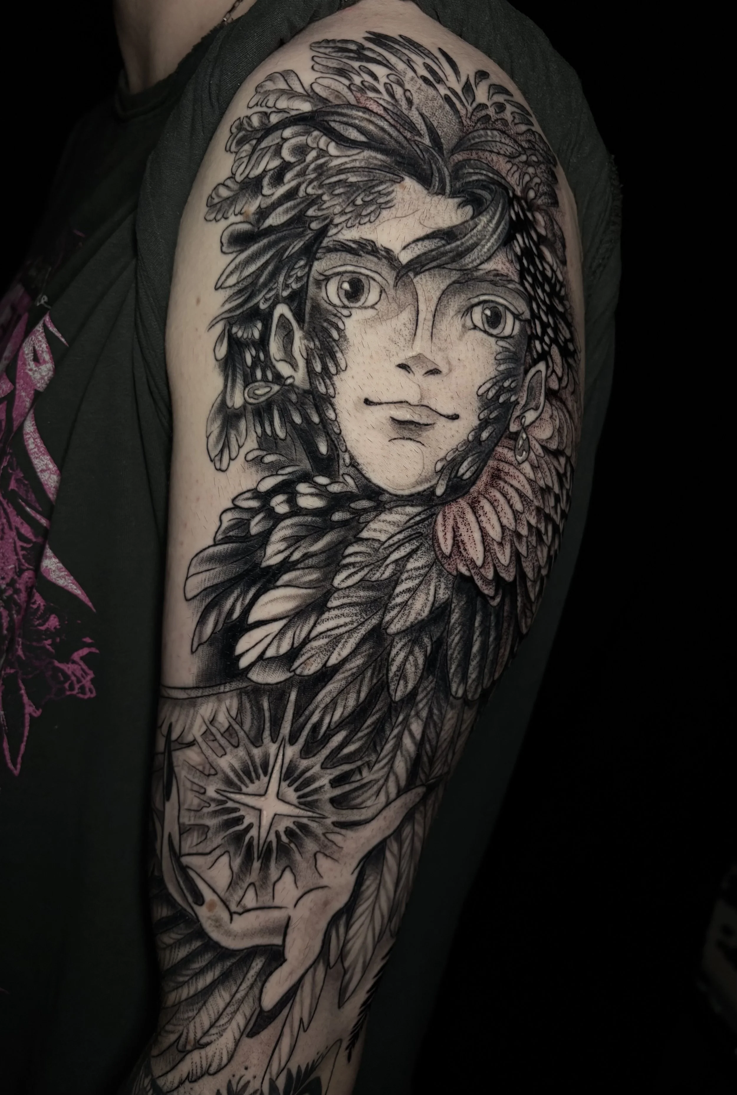 A detailed black and gray tattoo of a woman's face with hair blending into feathers, leaves, and a star-shaped symbol on the arm.
