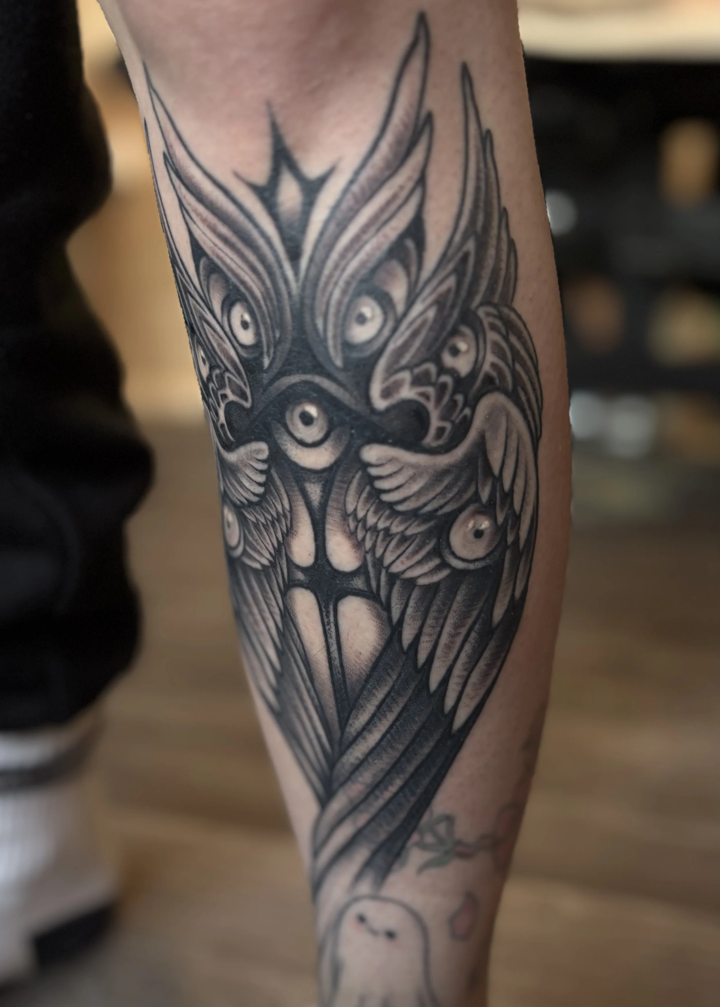 Black and gray tattoo on a person's arm depicting a bird with multiple eyes and intricate feather details.