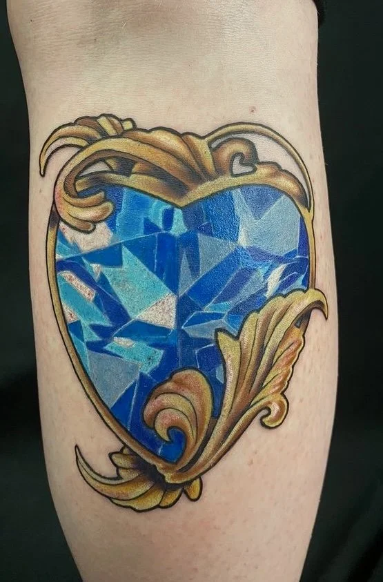 Tattoo of a large blue gemstone in an ornate gold frame on a person's arm.