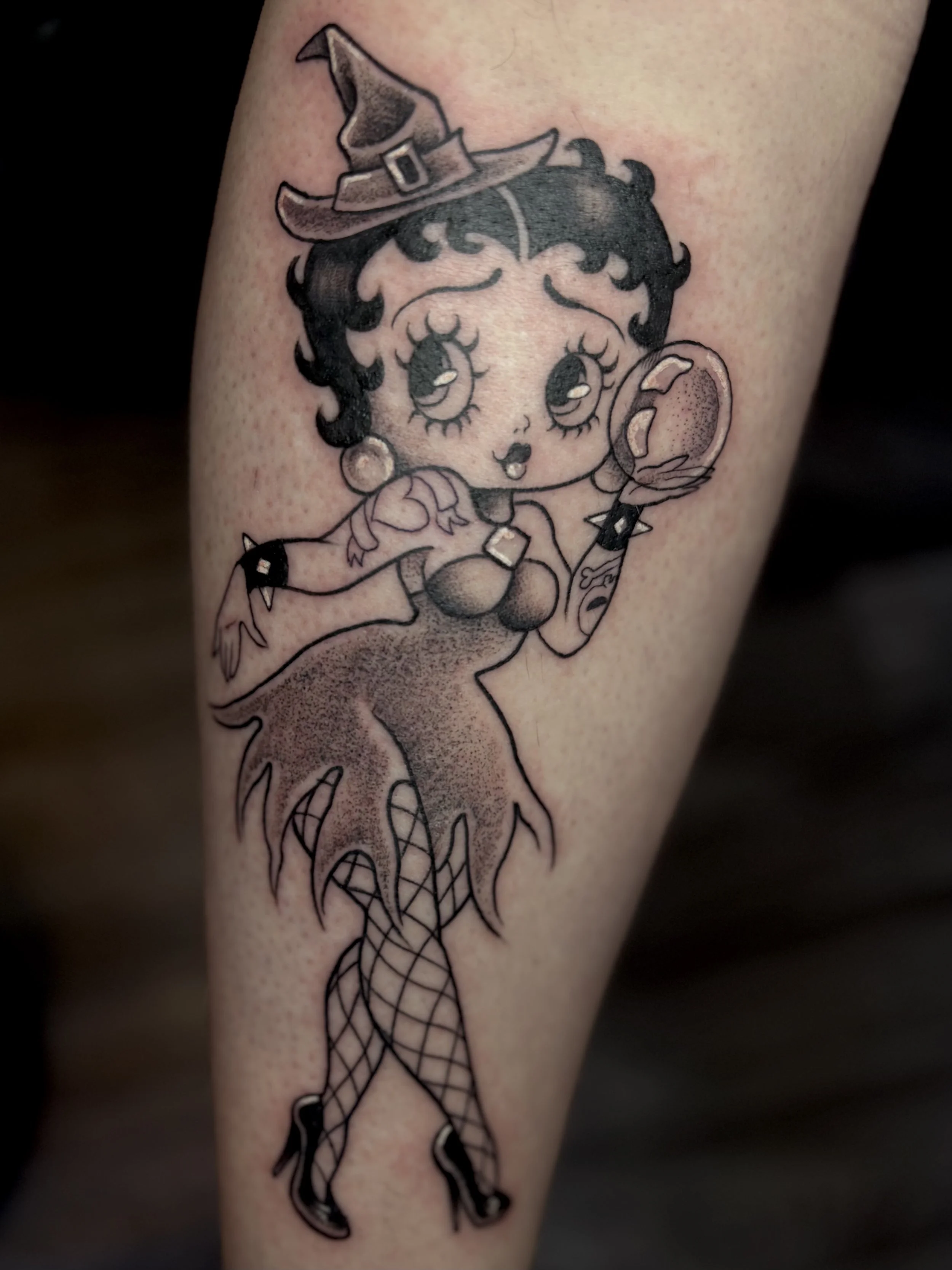 Tattoo of a cartoon woman dressed as a witch, holding a mirror, with a witch hat, fishnet stockings, and high heels.