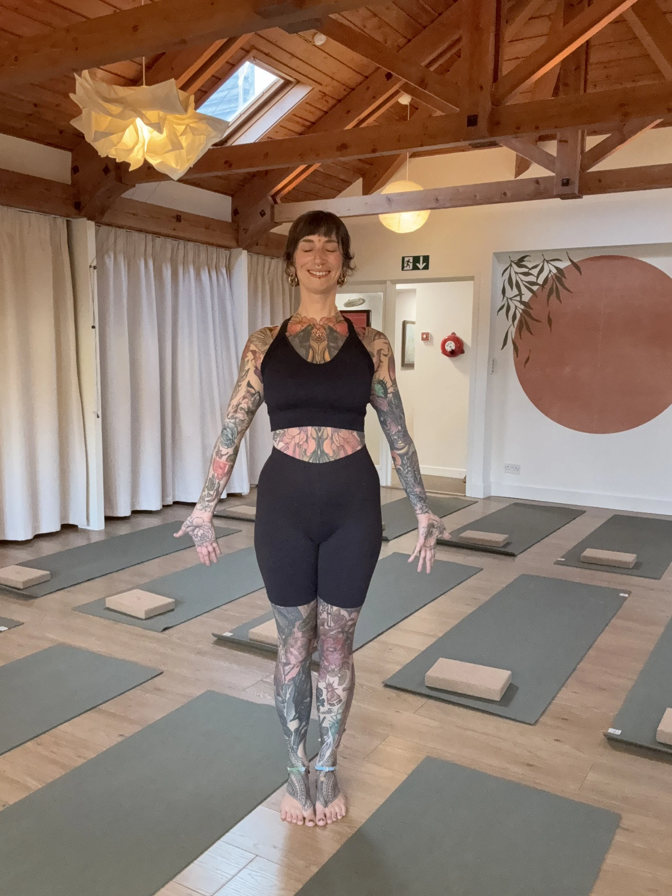 A woman with tattoos on her arms, legs, and chest, standing in a yoga studio with her eyes closed and smiling. The studio has wooden beams, yoga mats and blocks on the floor, and a large round artwork on the wall.