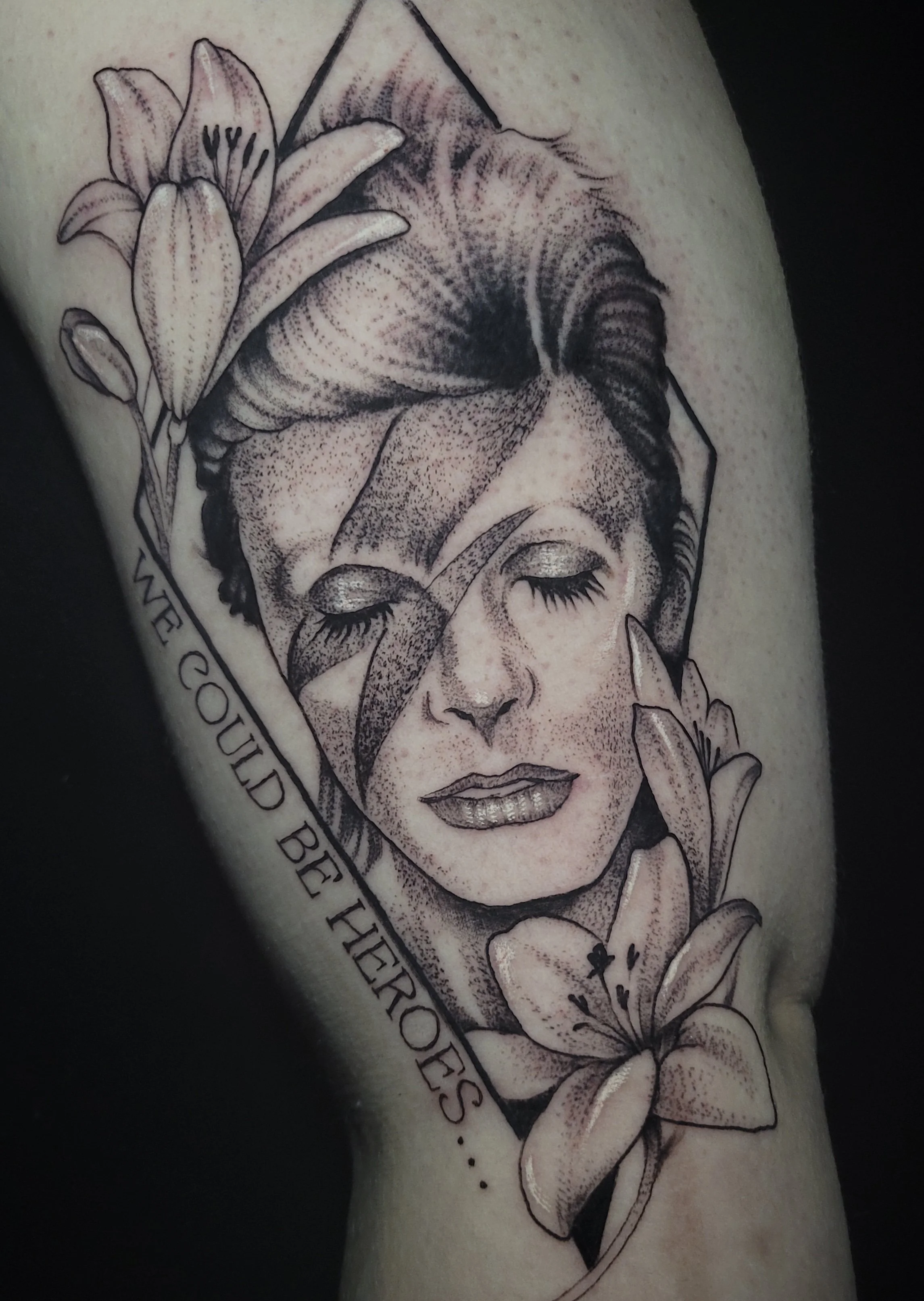 Tattoo of a woman's face with closed eyes, surrounded by lily flowers, and a quote that says 'We could be heroes...'