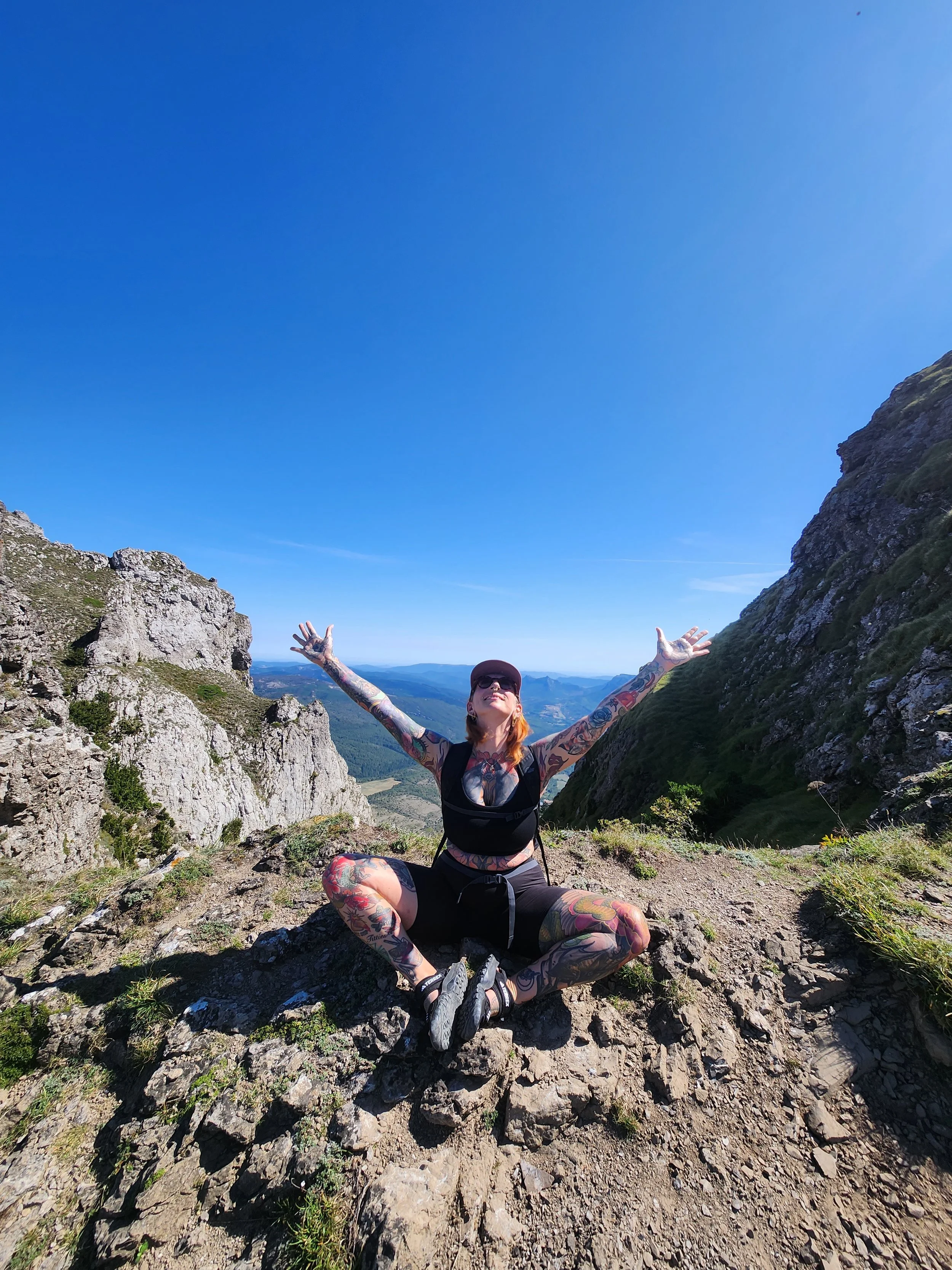 A woman with tattoos sitting cross-legged on a rocky mountain trail with arms raised, smiling, wearing athletic gear, in front of a scenic view of mountains and a clear blue sky.